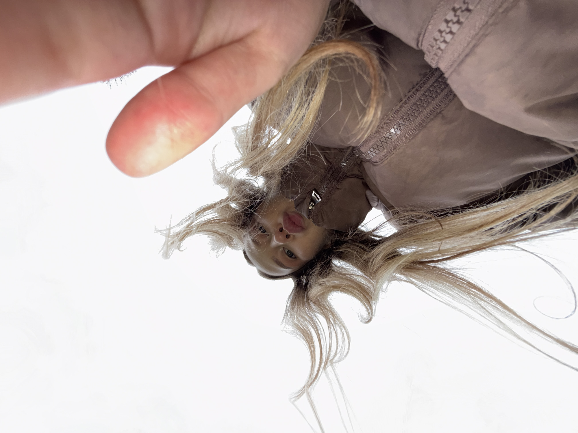 A low-angle, upward-facing shot shows a woman with blonde hair wearing a brown jacket. Part of a hand is visible in the top left corner of the frame. The background is a bright, washed-out white. The location is unspecified and not identifiable as Zhigalki, Russia.