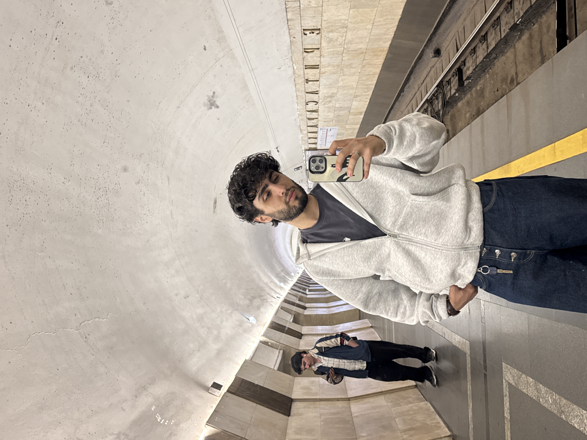 A man is taking a selfie in what appears to be a subway station. He is wearing a gray zip-up hoodie, a dark T-shirt, and dark blue jeans. He has dark curly hair and a beard. In his hand, he holds a smartphone with a light-colored case.
In the background, the station has a curved, arched ceiling and tiled walls with vertically aligned text. Another individual, wearing a patterned shirt and dark pants, is walking in the distance within the same station. The subway platform has a yellow safety line, and the tracks are visible in the background.