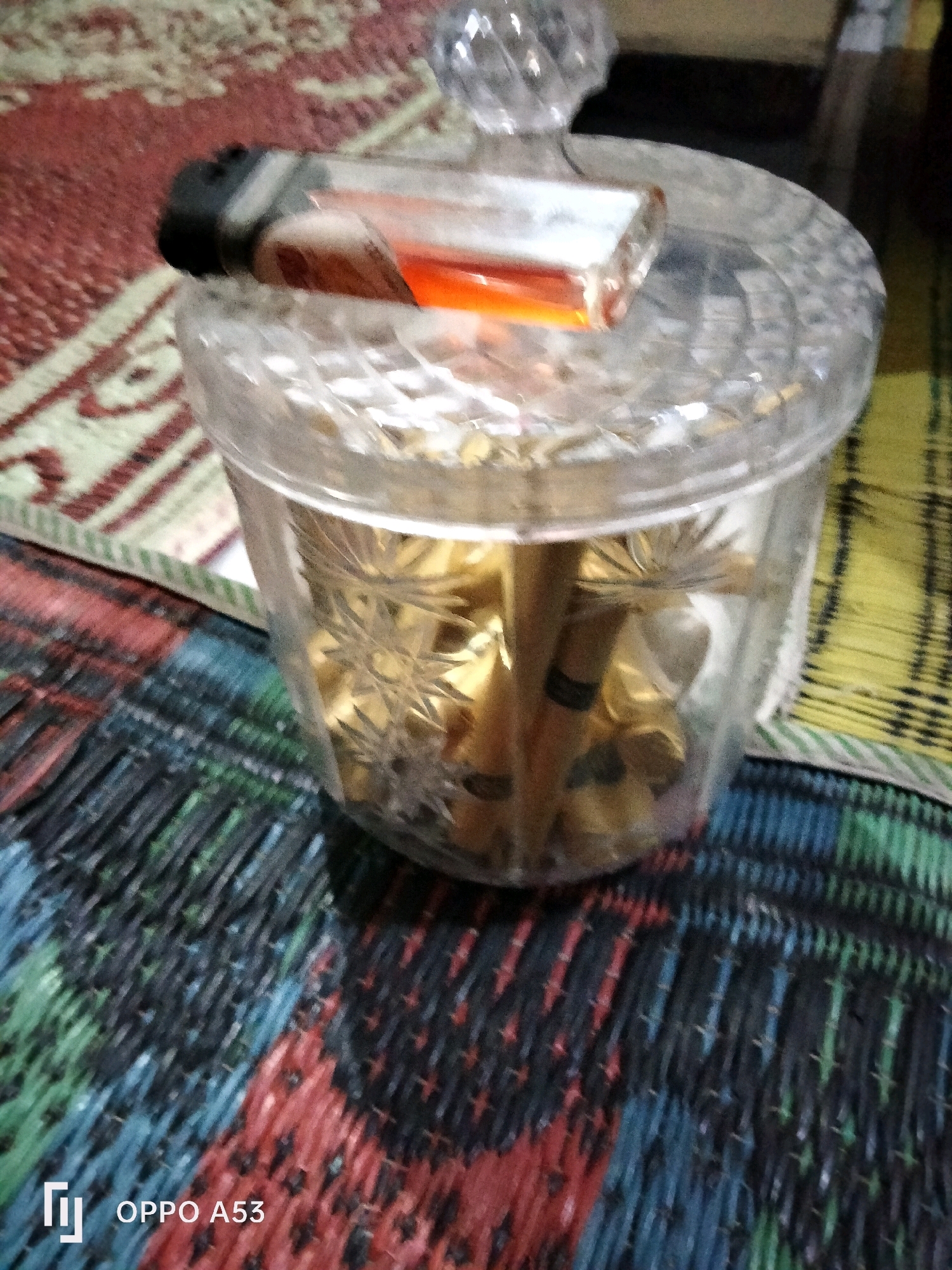 A close-up shot shows a glass jar filled with cigarettes, topped with a clear glass lid. A lighter, with its flame visible, rests on the lid. The jar is placed on a colorful woven mat, possibly a floor mat or a table runner, which appears to be on a larger patterned rug. The background is blurred, but suggests an indoor setting. The image contains a watermark in the lower left corner that reads "OPPO A53", indicating the device used for capture. The scene does not provide any specific visual cues to pinpoint its exact location within the city of Trenggalek, Indonesia, or any particular context of people, actions, or interactions.