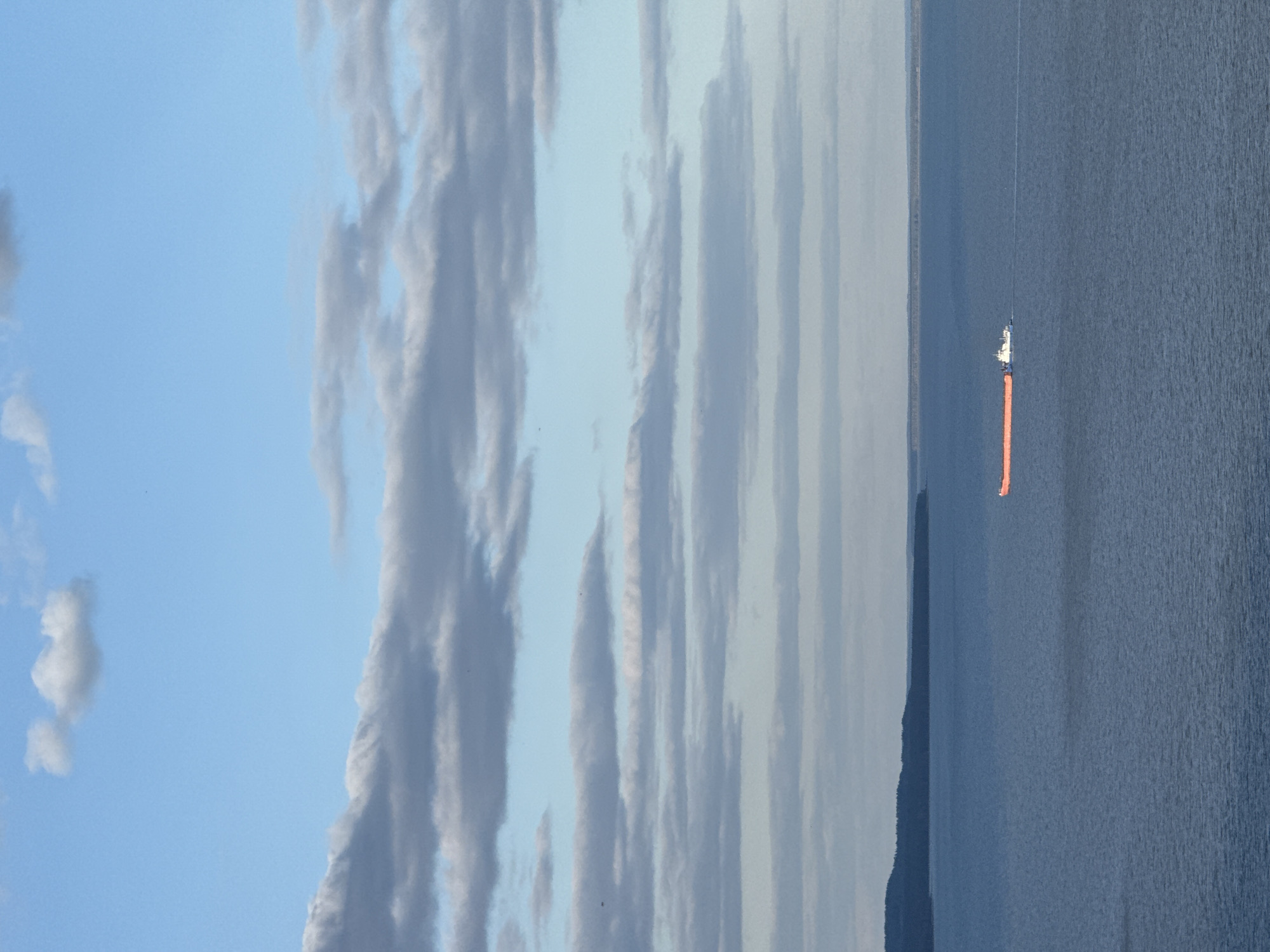 A vertical shot depicts a body of water, presumably a lake or ocean, occupying the right half of the frame. A solitary boat is visible on the water's surface, trailing an orange object behind it. Adjacent to the water, on the left, is a landmass, potentially a shoreline or an island. The sky above is filled with diverse cloud formations, ranging from wispy, linear clouds to larger, more textured cloud masses. The overall color palette is dominated by blues and grays, with a hint of orange from the object trailing the boat. The location is presumed to be near Galevo, Russia, based on the prompt.