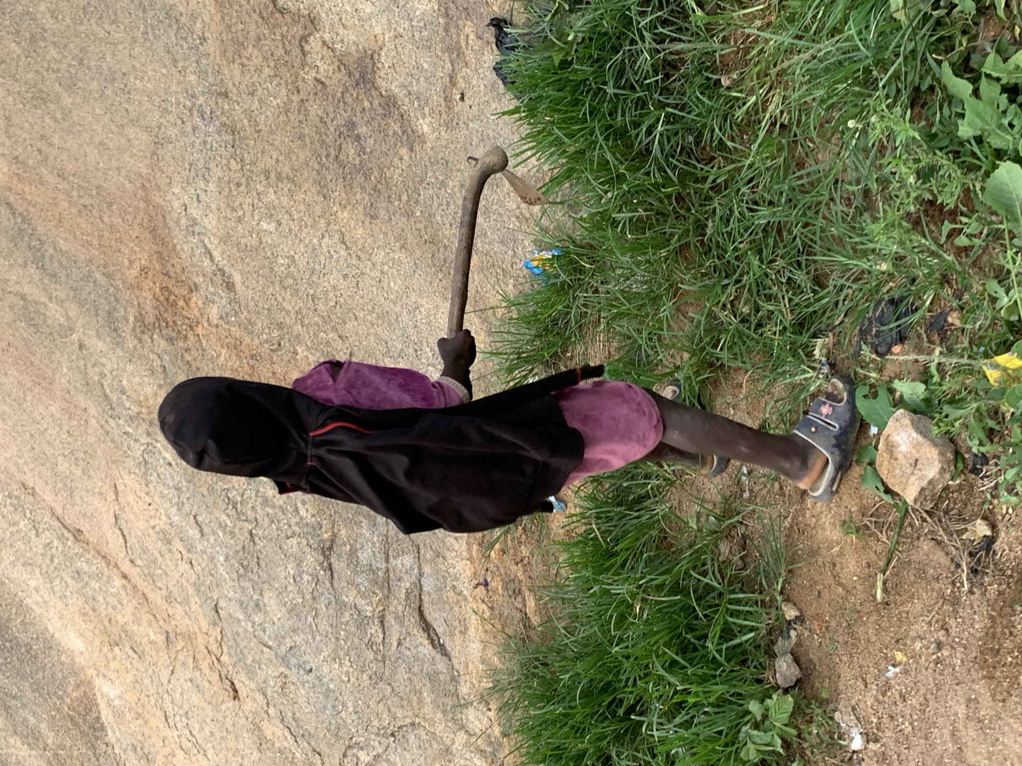 A person wearing a black hooded jacket over a purple dress and sandals walks outdoors with a hoe in their hand, carrying a black backpack. The person is standing in front of a large rock formation with green grass and dirt in the foreground. The scene appears to be located in Jos, Nigeria, as indicated by the presence of the rock formations characteristic of the region.