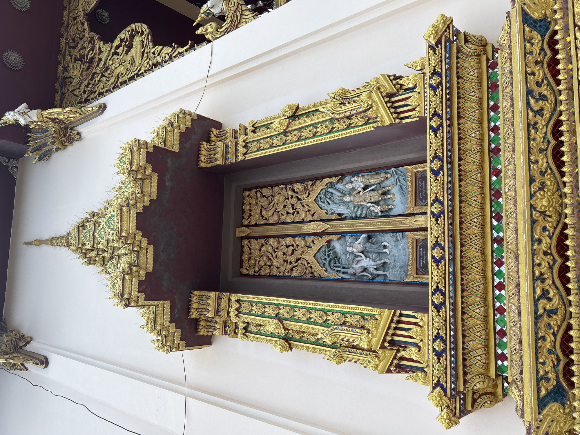 This is a low-angle, full shot of the exterior of a building in Ko Samui District, Thailand. The building's facade is predominantly white with ornate gold decorations. The main focus is a large, intricately carved golden doorway with a dark red archway above it. The doorway is flanked by elaborate gold pillars adorned with green accents. Below the doorway, there is a detailed golden platform with colorful mosaic tile inlays of blue, green, and red. The carvings on the door depict two scenes with figures. The overall impression is one of opulent, traditional Thai architecture.