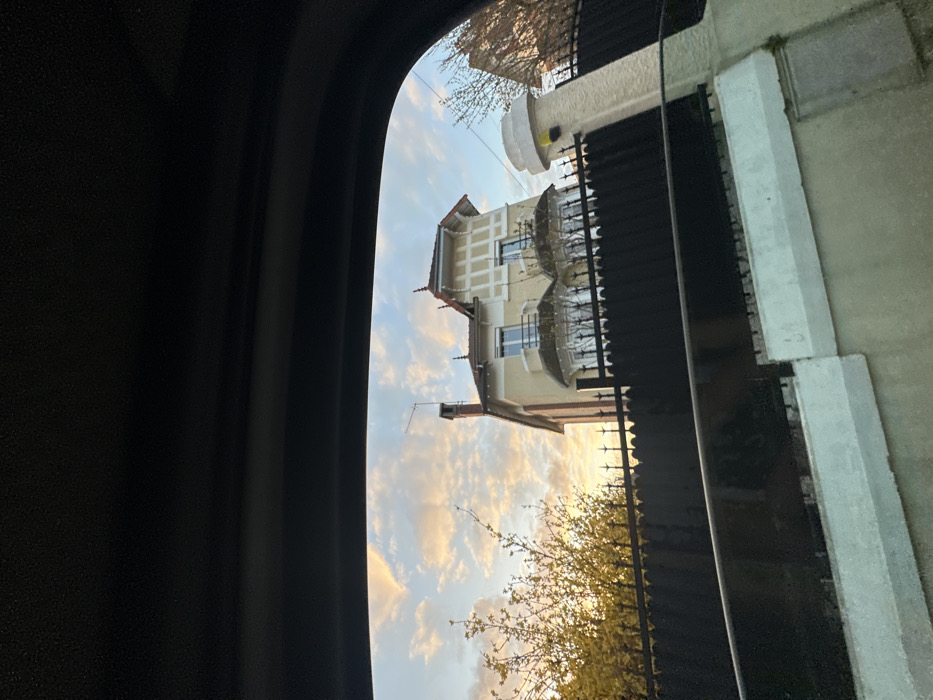 The image depicts a three-story residential building in Saulx-les-Chartreux, France, viewed from inside a vehicle, likely through a side window, with dark vehicle interior framing the top and bottom. The sky, characterized by scattered cirrus clouds illuminated with warm orange and pink hues, suggests either sunrise or sunset.
The primary subject is a cream-colored house featuring a steep dark-tiled hip roof with decorative finials. A prominent brick chimney with an antenna rises on the left side of the roofline. The facade includes multiple windows, with a notable two-story bay window structure centered on the first and second floors, featuring small, bare ornamental trees in front of the ground floor windows. Decorative white and cream timber framing patterns are visible on the gable ends and upper sections of the facade.
In the foreground, a dark metal fence with pointed pickets runs horizontally across the scene, partially obscuring the lower portion of the house. Below the fence, a light-colored stucco wall with a white concrete coping forms the property boundary. To the far right, a sturdy stucco pillar with a distinctive yellow security light or sensor cap is visible. On the left side of the frame, bright yellow-blossoming trees, likely forsythia, are silhouetted against the sky. No individuals are visible.