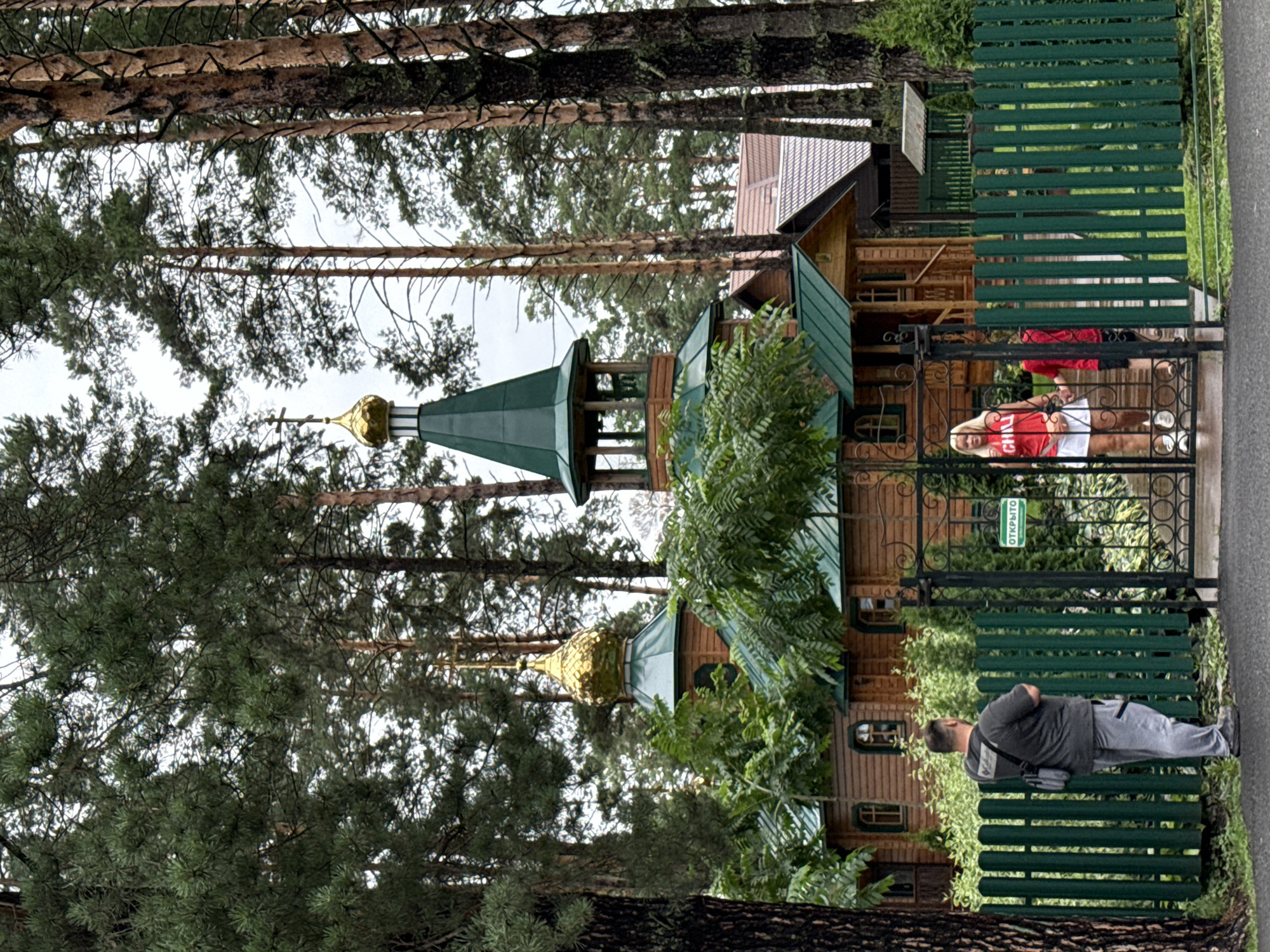 In Manzherok, Russia, a wooden Orthodox church with a green roof and golden domes is nestled among tall pine trees. A dark green metal fence with an ornate gate encloses the church grounds. Through the gate, two women and a child are visible. One woman, wearing a red t-shirt and white skirt, stands near the gate, while a child in a red shirt walks through it. A man in a gray t-shirt and pants stands outside the fence, looking towards the church. A green sign above the gate reads "ОТКРЫТО" (Open). The scene is set in a forested area with lush greenery.