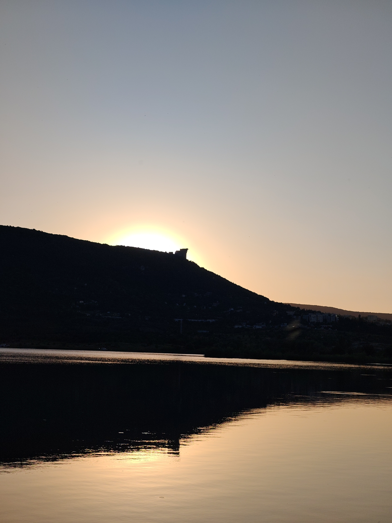 This is a vertical, wide-angle photograph of a sunset over a body of water, likely a lake or reservoir, with a silhouette of a hill and structures on its crest. The sun is partially obscured by the crest of the hill, casting a bright orange glow. The sky transitions from a pale blue at the top to a soft yellow-orange near the horizon. The hill is dark and silhouetted, with some faint lights visible on its slopes and on the crest, suggesting buildings or structures. The water in the foreground is dark, reflecting the muted colors of the sky and the silhouette of the hill, with the reflection of the sun creating a shimmering, golden path across its surface.
The location is identified as the area around the Shumen Plateau, near the city of Shumen, Bulgaria. The prominent structure on the hill's crest is likely the Monument "1300 Years of Bulgaria," a significant historical monument located on the plateau. The buildings visible on the slopes could be part of the residential areas or tourist facilities surrounding the plateau. The overall scene depicts a serene natural landscape at dusk, with human-made elements subtly integrated into the environment.