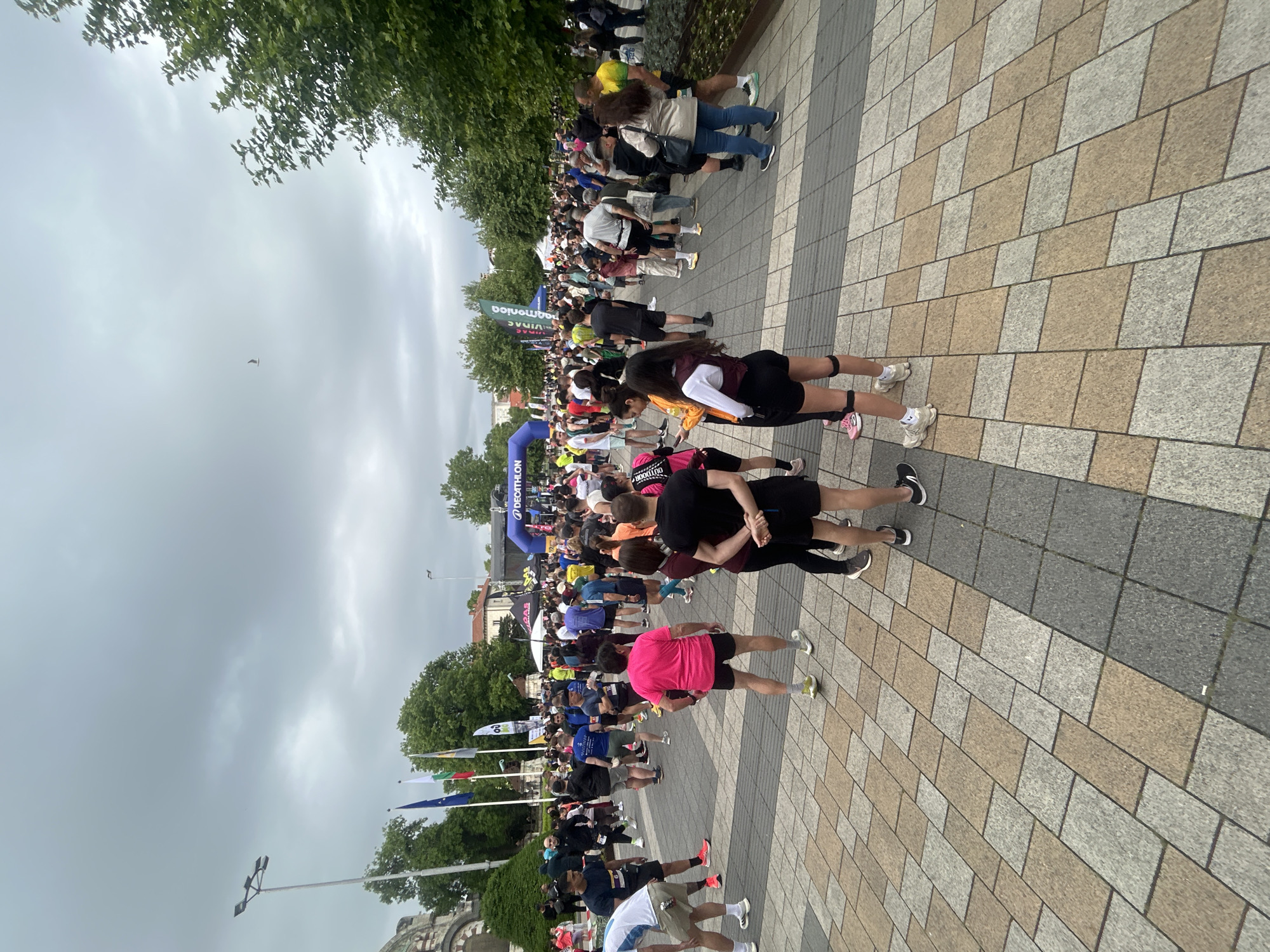 The image shows a large crowd of people gathered in what appears to be a public square in Pleven, Bulgaria. People are standing on a stone-paved surface, with some stretching or interacting with each other. Banners and flags are visible, suggesting a planned event or gathering, possibly a race or marathon, with an archway labeled "DECATHLON". Trees line the edges of the square. The sky is overcast, indicating cloudy weather.