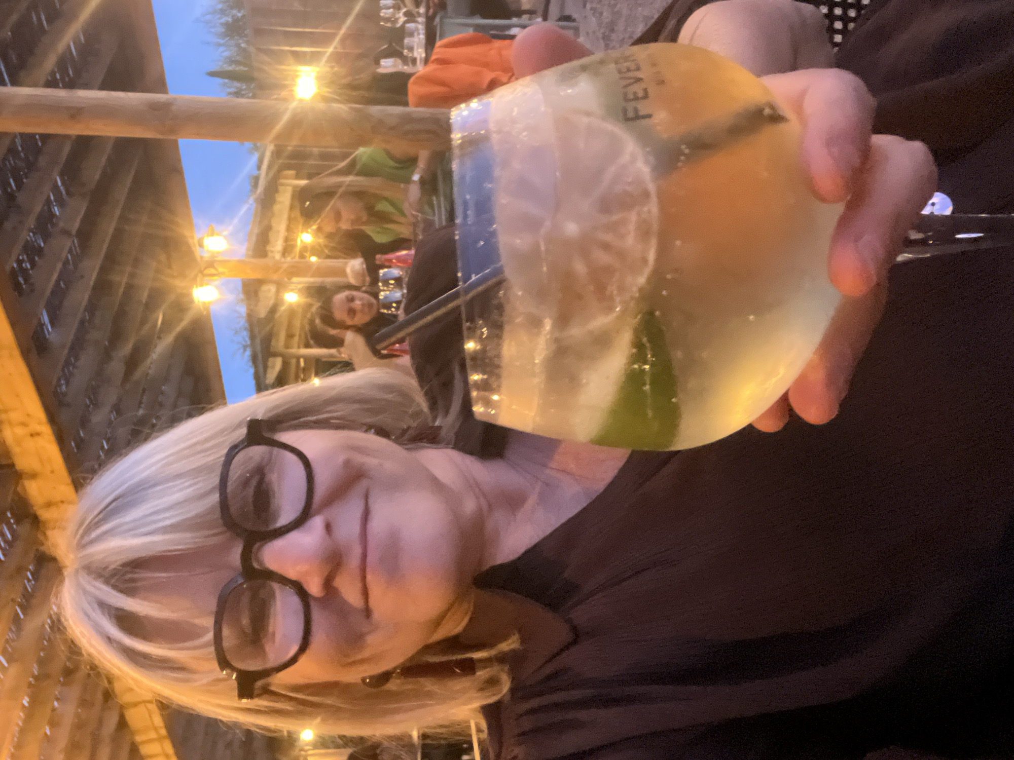 A woman with blonde hair and black glasses is holding a large cocktail glass with "Fever" on it. The cocktail has lime and orange slices and bubbles. The setting is a bar or restaurant in Andrézieux-Bouthéon, France, possibly outdoors under a wooden structure with hanging lights. Other people are seated at tables in the background.