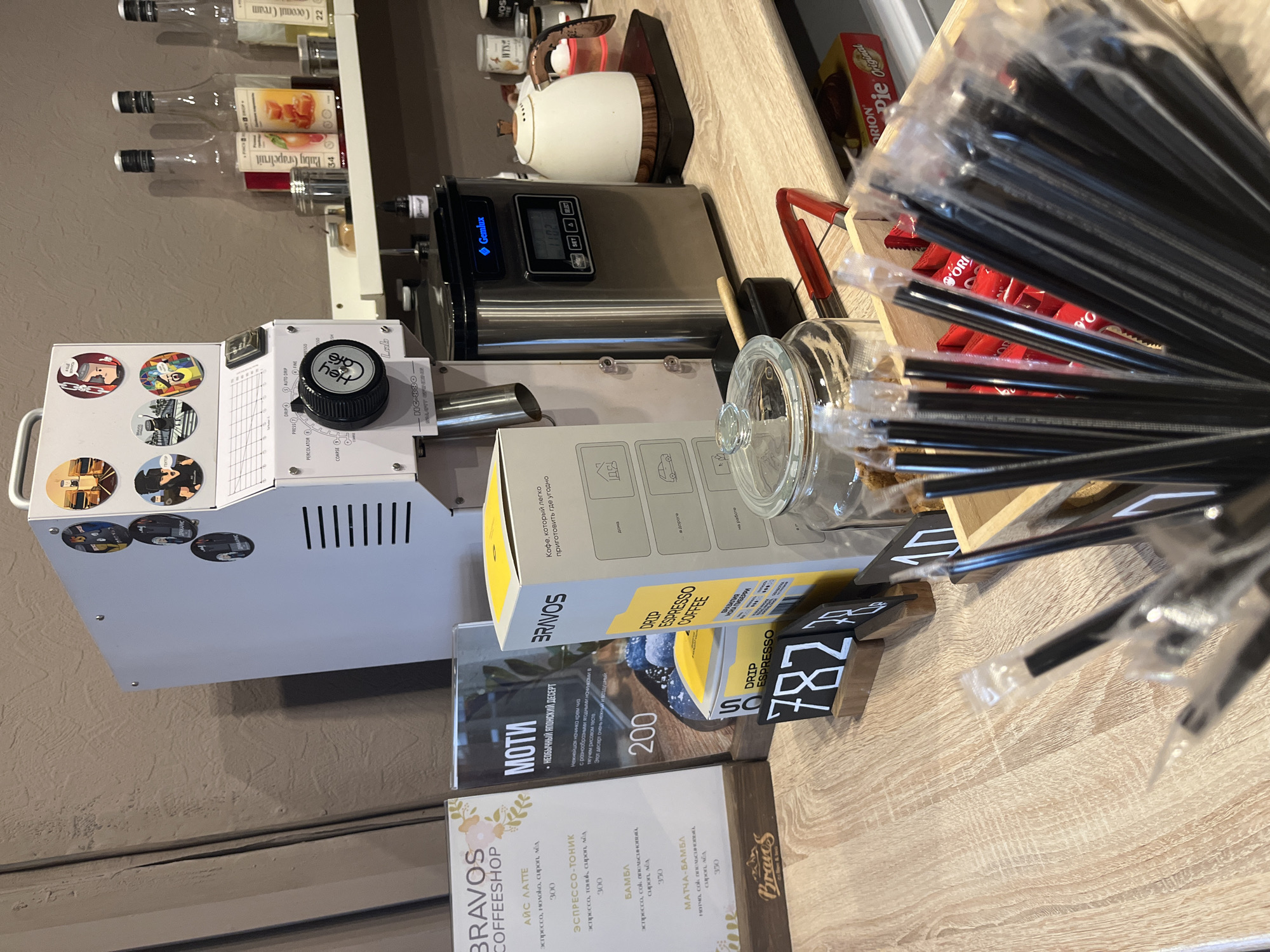 This image shows a coffee preparation area, potentially within a café or coffee shop, likely in Chaikovsky, Russia, as suggested by the brand name and Russian text on the promotional materials and menu.
Objects visible: A Heylo HC-880 coffee roaster, syrups, a white teapot, a Genlux appliance, a glass container, and a box of Brazos drip espresso coffee. A menu displays offerings like "ice latte", "espresso tonic", "Bambl" and "Matcha Bambl" in Russian, along with the price. There is also a sign advertising Japanese Mochi dessert. A container is filled with individually wrapped black plastic straws.