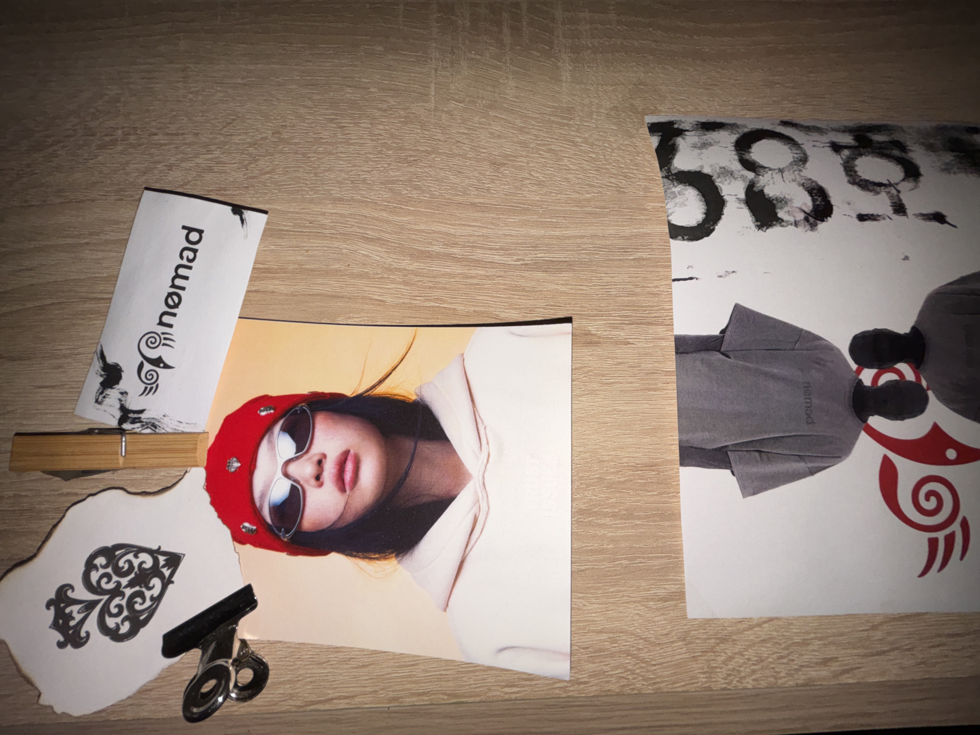 The image presents an overhead view of several papers and objects arranged on a light-colored wooden surface, likely a table or desk, illuminated by indoor lighting.
Central to the composition is a print depicting a stylized portrait of a person, seemingly a woman, from the shoulders up. She wears a red beanie and large reflective sunglasses, obscuring her eyes, with a neutral expression on her visible lips. She is dressed in a light-colored hooded top, set against a soft yellow background.
To the right, a larger, partially visible print is rotated ninety degrees clockwise. This print shows the lower torso of a figure wearing a grey t-shirt that has "nomad" printed on it. Below this, a bold red tribal-style design with a central swirl and claw-like extensions is prominent. The top portion of this rotated print features abstract black numbers or symbols, possibly "588," scrawled on a white background.
Other items include a small, white rectangular strip of paper positioned upside down, displaying the text "nomad" with a distinctive logo incorporating tribal elements. A wooden clothespeg-style clip lies near this paper. Additionally, an irregularly shaped piece of white paper, with edges appearing burnt or charred, features an elaborate black spade symbol. A black metal binder clip rests on this spade-emblazoned paper.
No specific activity is depicted; rather, the arrangement appears to be a display or collection of themed materials. The recurring "nomad" text and tribal motifs suggest a common brand or aesthetic across the prints. The scene provides no direct geographical cues beyond an indoor setting, despite the specified location context of Troyan, Bulgaria.