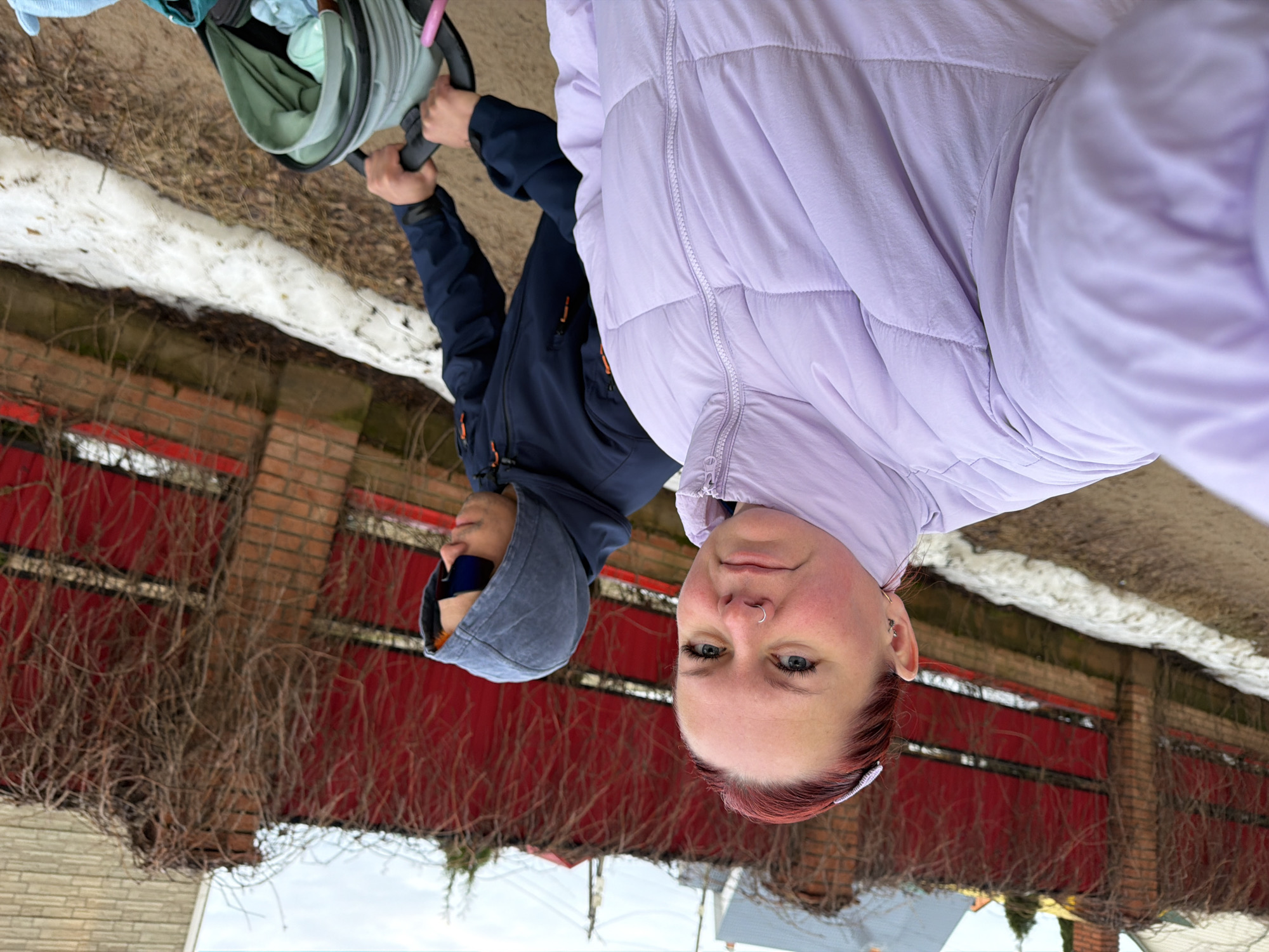 The image depicts two people, one holding a baby stroller, in an outdoor setting. A woman with reddish-purple hair and a light purple puffer jacket is facing the camera. Beside her, a person in a dark blue hooded jacket and sunglasses is pushing a baby stroller. The background includes a red brick wall partially covered with dry vines, patches of snow, and glimpses of buildings. The location appears to be the city of Chaikovsky, Russia, based on contextual clues such as architecture and typical winter landscape.