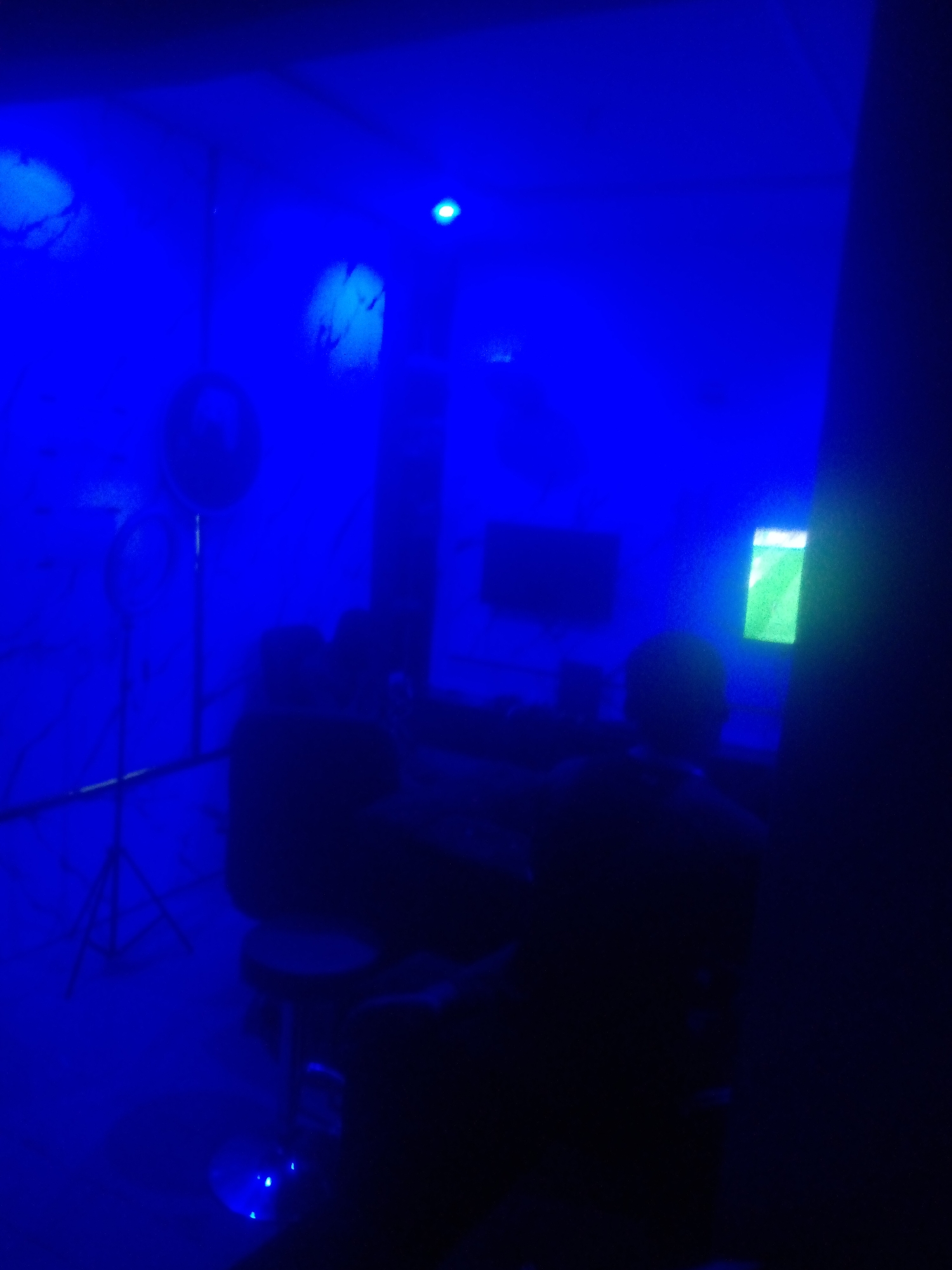 In a dimly lit room illuminated by a dominant blue light, a group of individuals appear to be gathered around a table, possibly engaged in gaming or a similar activity. The room's ambiance is further enhanced by what seems to be a professional ring light setup visible on the left side of the image, suggesting a potential recording or streaming session. A television screen displaying a green graphic is mounted on the far wall, and a single stool is positioned in the foreground, under a spotlight. The scene is set in Jalingo, Nigeria.