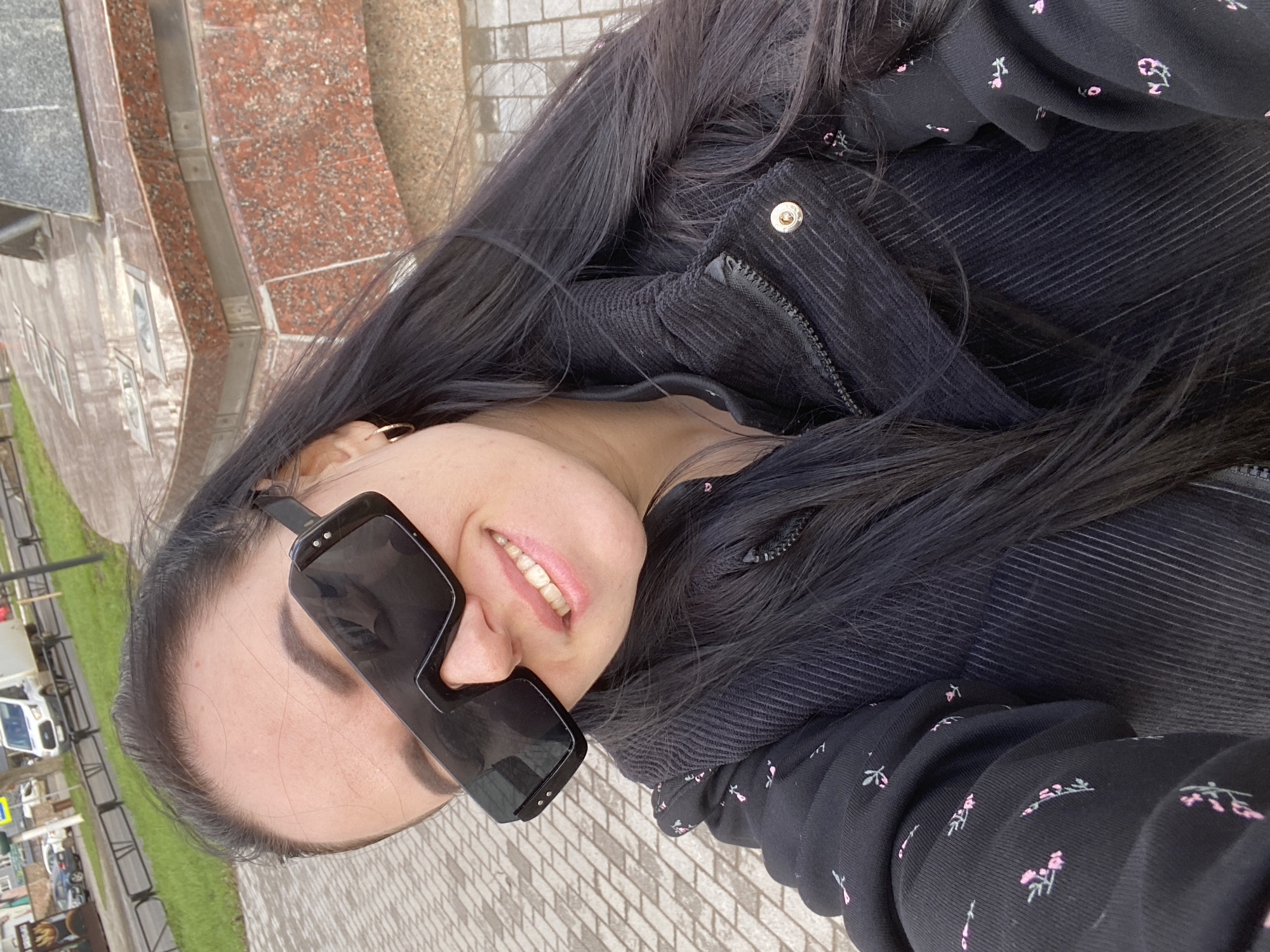 The image is a close-up selfie featuring a young woman taken outdoors in Chaikovsky, Russia. The woman, with long, dark hair, is smiling broadly, revealing her teeth. She is wearing large, black, rectangular sunglasses that cover a significant portion of her face. A small gold hoop earring is visible on her right ear. Her attire includes a dark, likely black, corduroy jacket with a visible zipper and a gold-colored snap button near the collar. Beneath or as part of the jacket, a dark fabric patterned with small pink floral or leafy motifs can be seen.
The background depicts an urban environment. In the immediate foreground, a light-colored brick pavement is visible. To the woman's left, there is a grassy strip and a street with parked vehicles, including a white van, and some metal barriers. To her right and extending into the upper background, a building with a facade of reddish-brown polished granite or marble and some grey stone is present. There appear to be unreadable white characters or designs on plaques affixed to this building's facade. The scene is illuminated by soft, diffused daylight, suggesting an overcast sky. The woman's expression indicates a cheerful or contented mood.