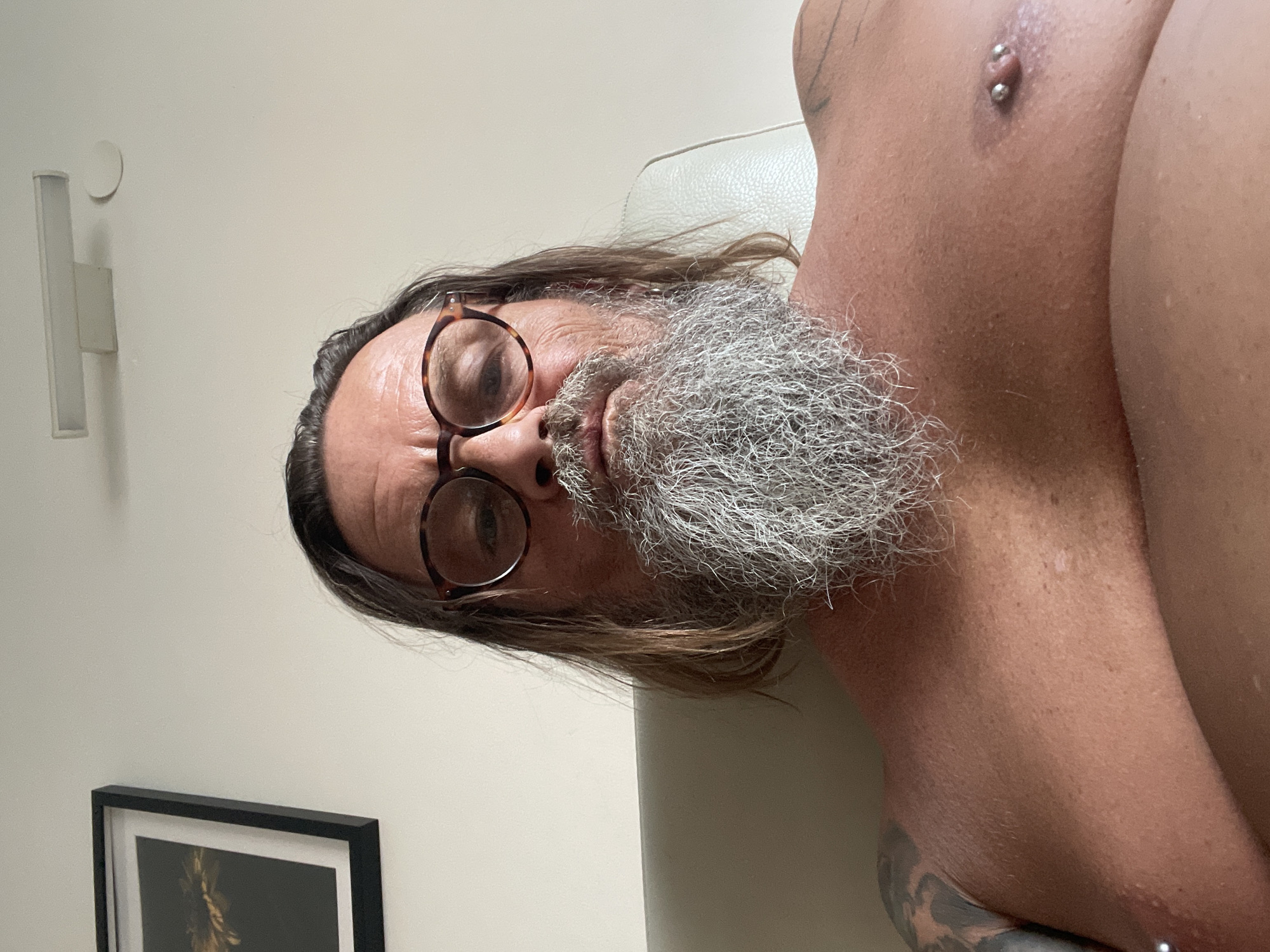 The image shows a close-up, forward-facing, selfie-style shot of a shirtless man with a long, graying beard and shoulder-length brown hair. He is wearing eyeglasses with tortoiseshell frames and has a piercing on his right nipple. He has tattoos on his left shoulder and upper arm. The man is seated on a light-colored couch. In the background, a framed picture hangs on a light-colored wall, and a modern light fixture is mounted on the ceiling. The environment appears to be indoors, likely a residential setting, and is not definitively identifiable as being in Netanya, Israel, as no specific landmarks or indicators of that location are visible.