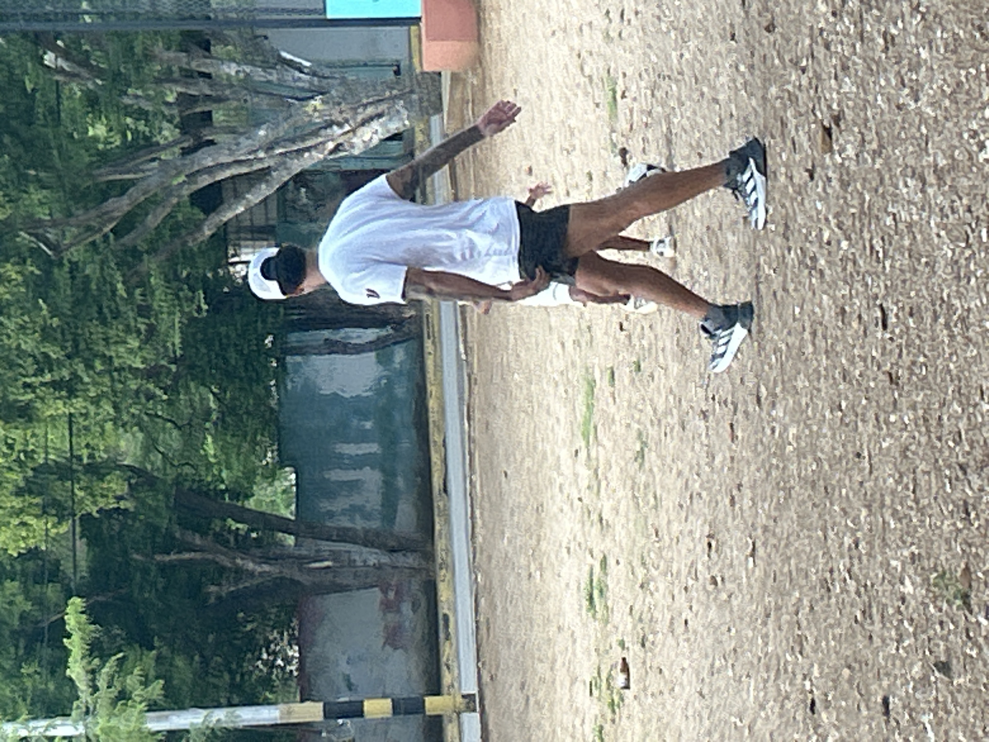 A man wearing a white t-shirt, black shorts, a white baseball cap, and athletic shoes walks across a sandy, outdoor area. He has tattoos on his arms. Another person, partially visible, stands behind him, also wearing athletic shoes. The background features trees with lush green foliage, a grey wall with some red markings, and a yellow and black striped pole. The ground is covered with sand and scattered small stones. The scene appears to be a public space, possibly a park or sports field, in Santa Ana, Venezuela.