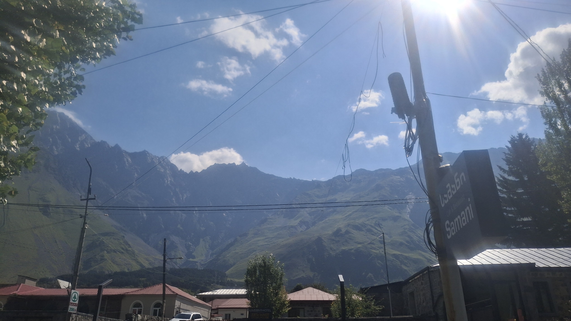 The image shows a street view in Stepantsminda, Georgia, with a prominent mountain range in the background. In the foreground, a utility pole stands to the right, adorned with a sign that reads "Samani" in English and also in Georgian script. The sign also indicates "Cafe & Restaurant". Various wires are attached to the pole and extend across the frame, intersecting with the sky and the mountain scenery.
On the left side of the image, tree branches with green leaves frame the upper left corner. Several houses with red roofs are visible in the mid-ground, with the mountain slopes rising behind them. The mountains are covered in a mix of green vegetation and rocky outcrops. The sky is bright blue with scattered white clouds, and the sun is shining intensely, causing lens flare in the upper right quadrant of the image. The overall scene suggests a rural or semi-urban setting at the foothills of a significant mountain range, likely in the Caucasus region.