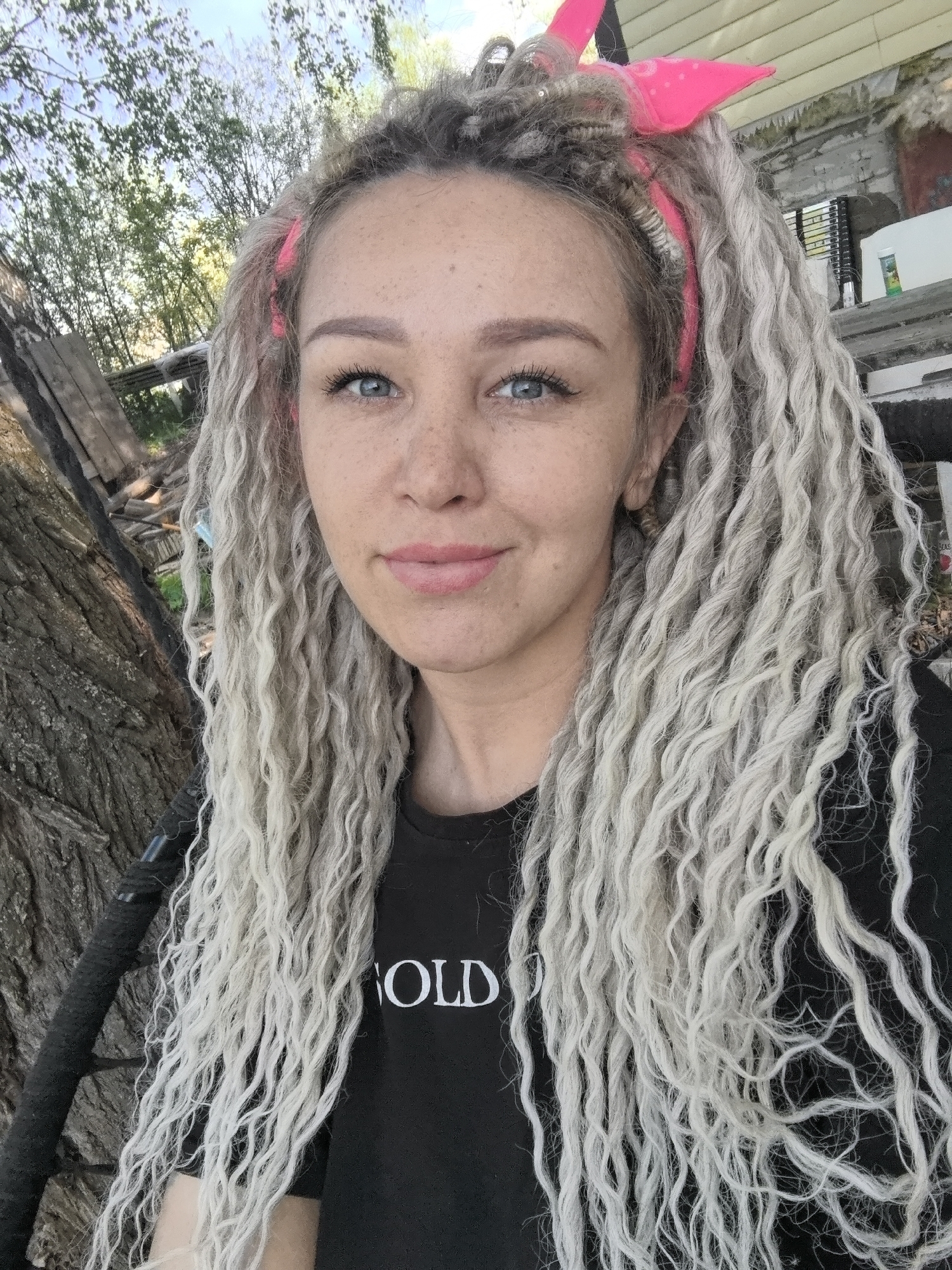 A young adult female with fair skin, freckles, and light blue eyes is centered in the frame. She has long, curly, silver-grey hair, styled in what appears to be dreadlocks, held back by a pink headband with a tied detail. She is wearing a black t-shirt with a white word that is partially visible.
She is positioned outdoors, with a tree trunk visible to the left side of the frame. The background shows foliage, suggesting a park or garden setting. The image has a slightly overcast appearance. The location, Noviy, Russia, is not discernible from the image.