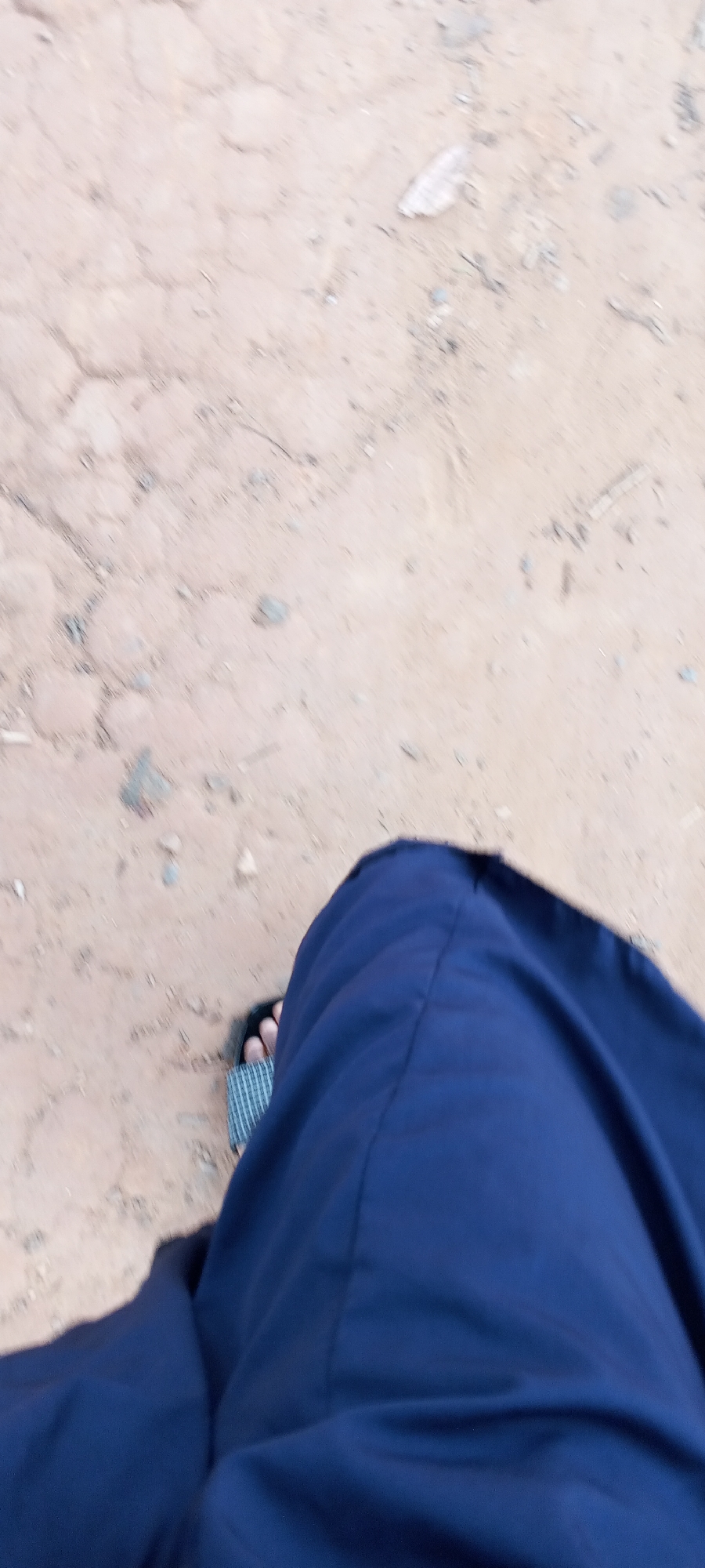 The image shows a ground-level view of a person wearing a blue garment and sandals. The background consists of sandy or dirt-covered ground with scattered pebbles and debris. The location is unspecified, but the question assumes it is in the city of Keffi, Nigeria. The primary subject is the lower portion of a person's body, with the focus on their clothing and footwear. No specific actions or interactions are discernible.