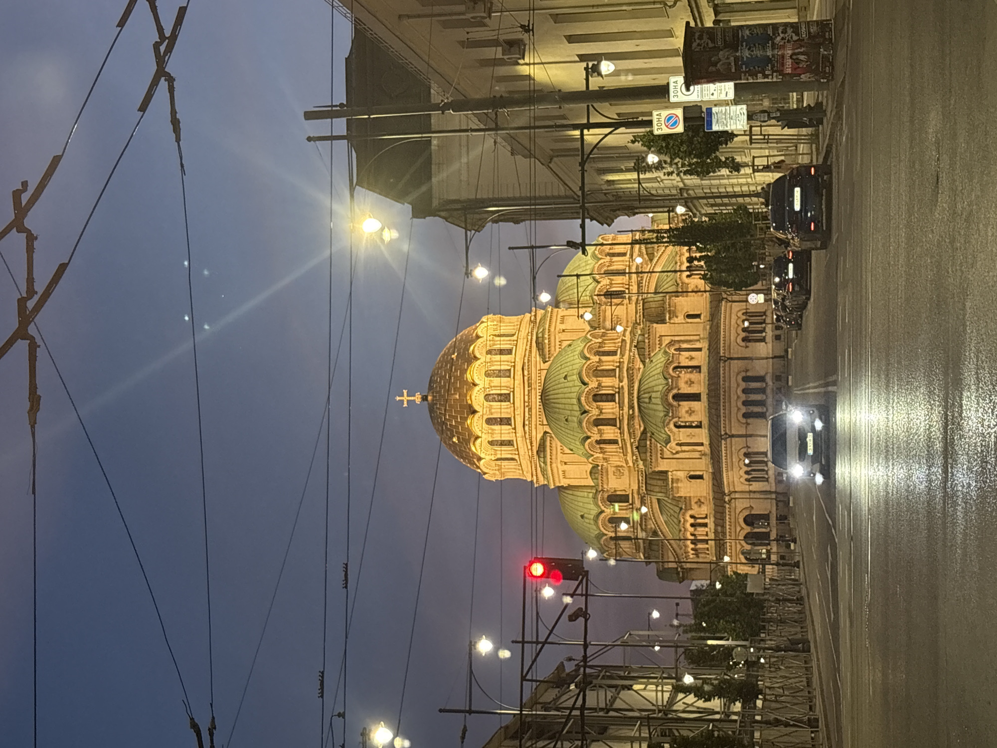 The image shows a street scene at dusk with the Alexander Nevsky Cathedral in Sofia, Bulgaria, illuminated in the background. The cathedral features a prominent golden dome topped with a cross and several green domes. The street is paved and wet, reflecting the headlights of a car driving towards the viewer. Several other cars are parked along the right side of the street. Overhead, a network of electrical wires is visible against the twilight sky. Streetlights are illuminated, casting a warm glow on the surroundings. The overall atmosphere is calm and serene.