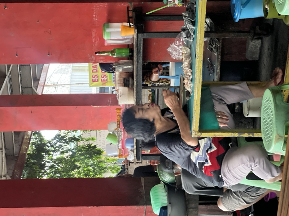 An older woman with short, dark hair is seated at a yellow-topped, green-framed metal table in an outdoor or semi-enclosed eating area in Nganguk, Indonesia. She wears a black short-sleeved top with a white and red patterned design on the sleeves and light-colored pants, along with red flip-flops. She holds a spoon to her mouth, actively eating from a dark bowl positioned in front of her. Her lips are pursed. On the table to her right are several small, light-colored, irregular objects, possibly peeled garlic or small food items, alongside a crumpled dark cloth. She wears a gold ring on her right ring finger.

Behind her, a person wearing a dark gray shirt and a dark cap is partially visible, seated on a light green plastic stool, facing away from the camera. The setting features prominent red structural pillars. To the right of the main subject, a metal shelving unit holds various items, including a green glass bottle, clear plastic containers, a white pitcher, an orange cup, and shakers. A yellow banner with "ES DAY..." and "ES..." (likely indicating "ice drinks") is visible in the background. Green foliage is visible beyond the pillars on the left, while other kitchenware, including a green plastic colander, is visible on a lower shelf to the left of the main subject. The floor is a concrete or dirt surface, with a blue bucket and other items visible beneath the table.