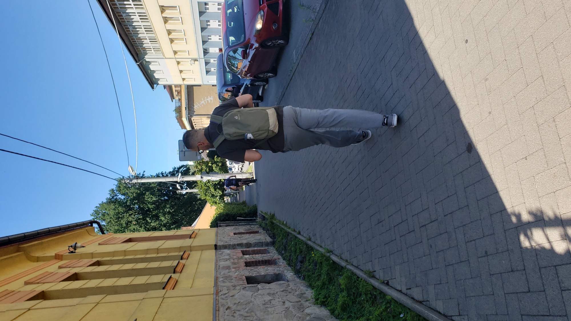 A man with short dark hair and glasses, wearing a dark polo shirt, grey trousers, and black and white sneakers, walks away from the camera on a paved sidewalk. He has a light green backpack on. He is on a sidewalk in front of a yellow building with stone accents and barred windows. Across the street, there are parked cars and buildings, including a cream-colored building with a grey roof and white-framed windows. The sky is clear and blue. The scene appears to be on a street in Prešov, Slovakia.
