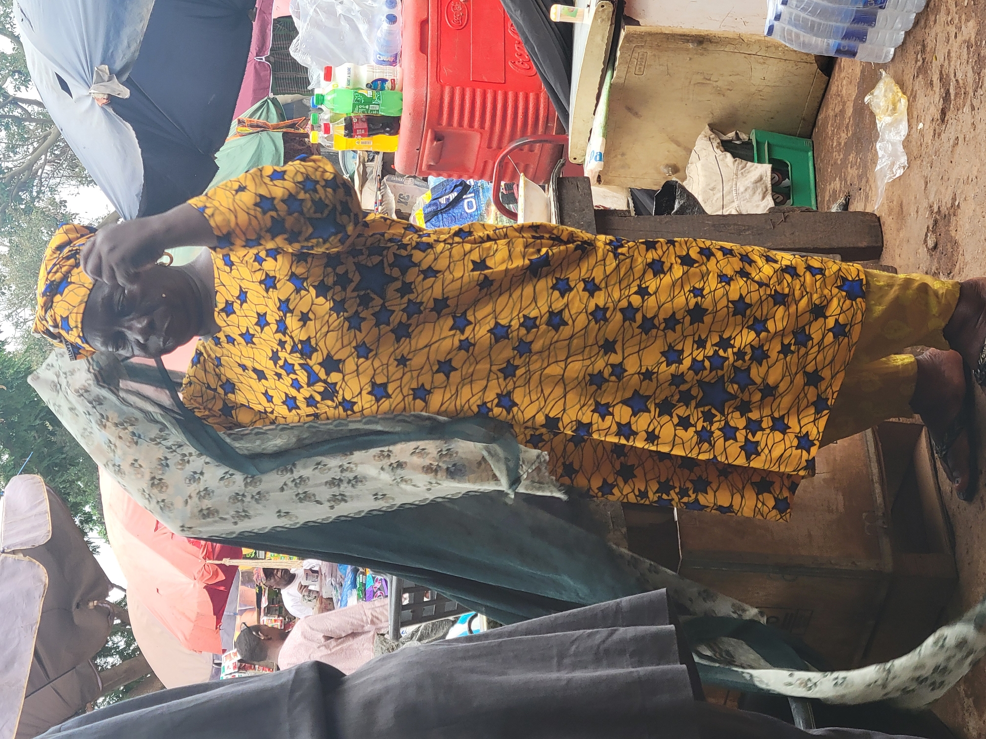 An elderly woman with dark skin stands in what appears to be a market stall in Abuja, Nigeria. She is wearing a bright yellow and blue star-patterned dress with a matching headwrap. She is adjusting her head covering with her right hand. Her facial expression is obscured by the angle of the camera and her hand.
The stall contains a variety of items, including plastic water bottles, a red cooler, and other assorted goods, all situated on a makeshift countertop. Numerous fabrics hang in the foreground, suggesting she might be selling textiles. The stall is under umbrellas. Other people are visible in the background. The stall appears to be on a dirt surface.