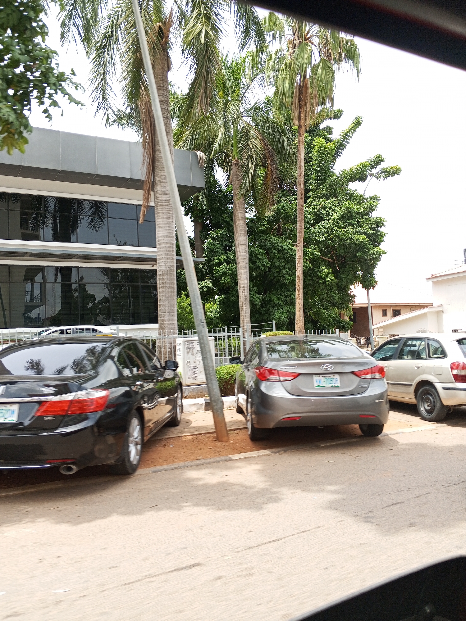 The image shows three parked cars on a street in Abuja, Nigeria. The cars are, from left to right: a black Honda Accord, a gray Hyundai Elantra, and a light-colored hatchback. They are parked next to a building with dark windows and palm trees in front. A white fence separates the building from the cars. The street is paved and has a curb. There are green trees in the background. A utility pole is visible near the gray car.