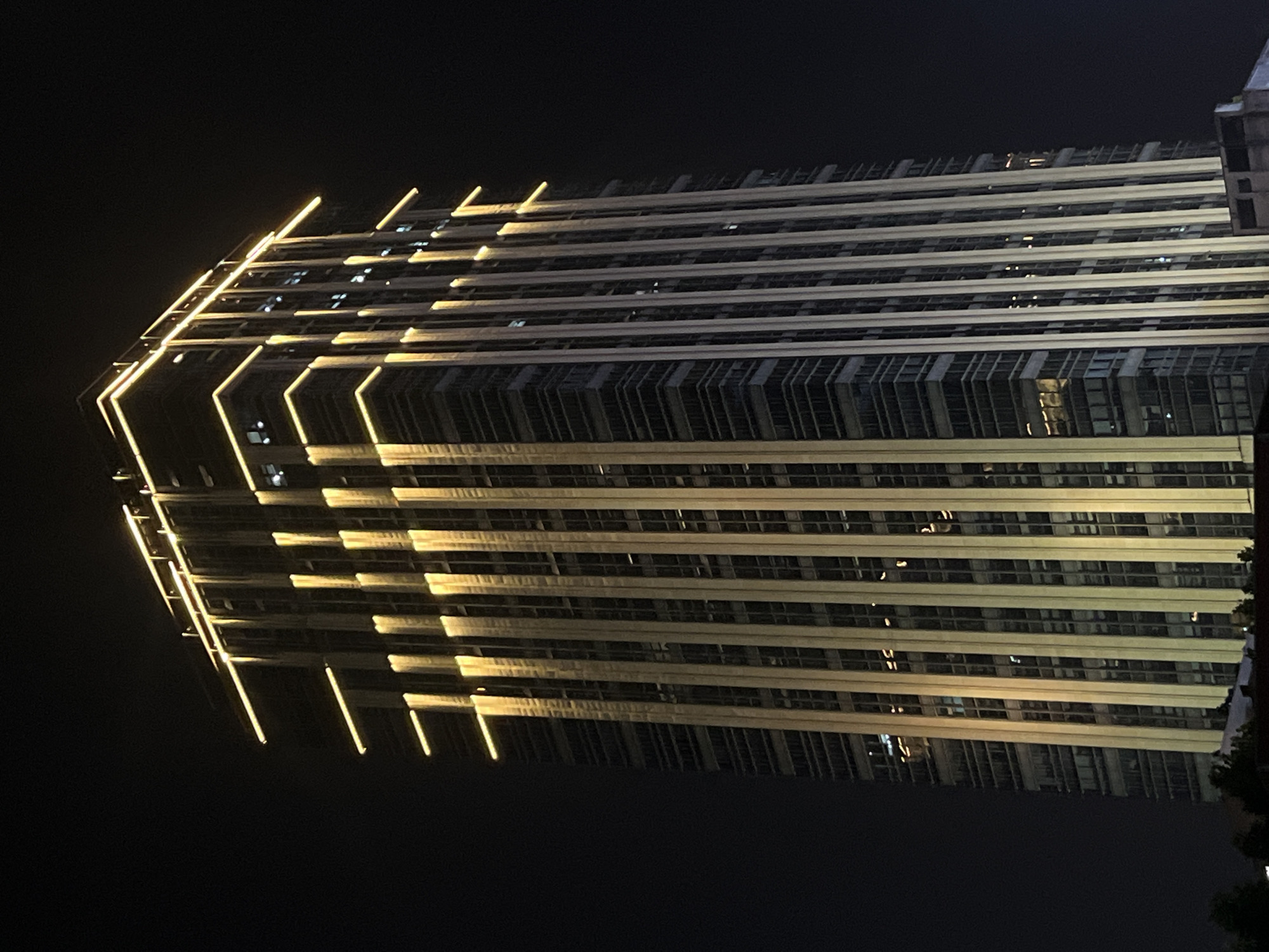 The image presents a tall, modern skyscraper illuminated at night in Yuzhong District, China. The building, rectangular in form, dominates the frame against a completely dark night sky. Its facade is composed primarily of glass windows and darker structural elements. The architecture is significantly highlighted by extensive golden-yellow lighting. Linear light strips precisely outline the building's roofline and at least three distinct upper horizontal tiers. Additionally, multiple prominent vertical light strips ascend portions of the facade, creating a series of illuminated columns. Scattered interior lights are visible through some of the building's windows. No individuals or specific actions are discernible within the frame. Minimal dark shapes, possibly representing other structures or foliage, are faintly visible at the very bottom edge of the image, suggesting an urban environment.