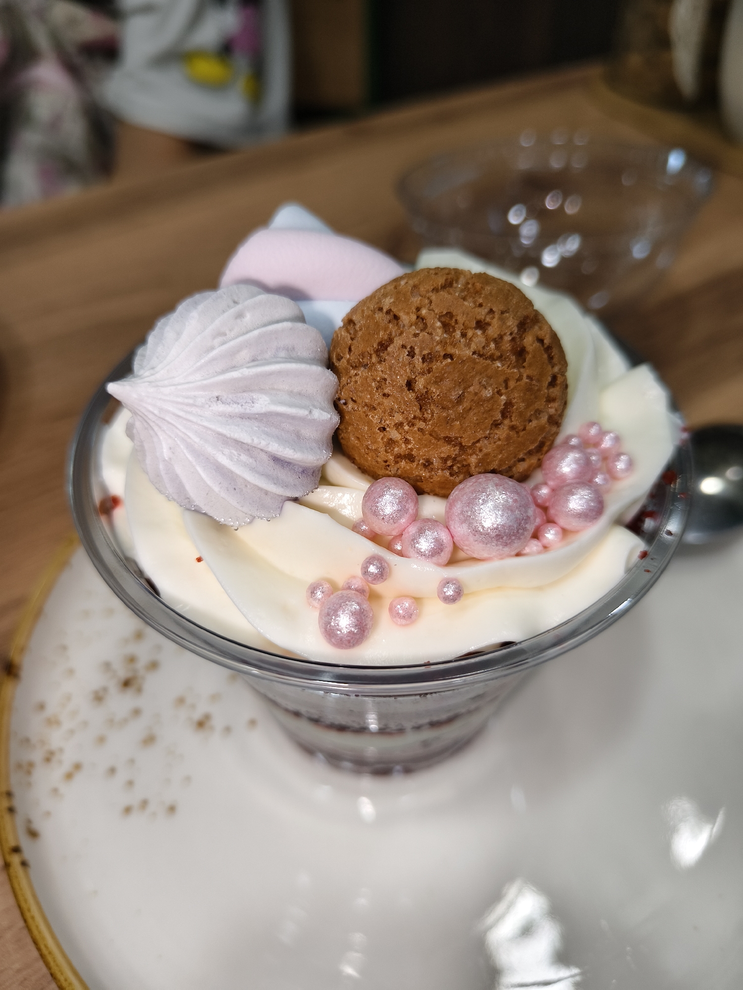 A clear glass dessert cup sits on a white saucer with speckles. The cup contains layers of dark red cake, white cream, and is topped with a swirl of white cream. A light purple and white meringue is placed on the left side of the cream. A small, brown, round cookie sits on the right side of the cream. Pink, shimmery edible pearls of various sizes are scattered on top of the cream and around the cookie. In the blurred background, a person wearing a white top with a yellow cartoon character is visible. Another blurred object, possibly a glass bowl with a reflective surface, is in the upper right background. The scene is likely indoors, with the lighting suggesting a well-lit environment.