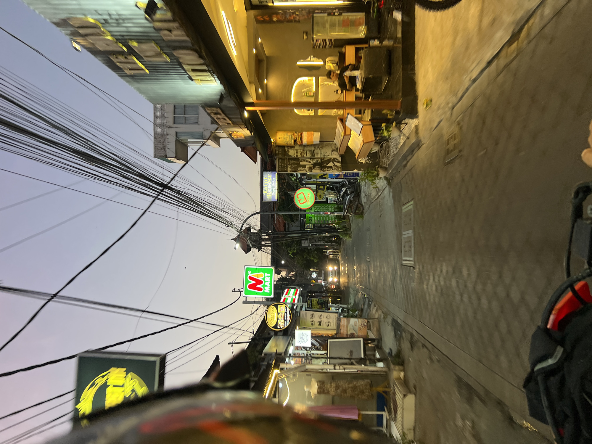 An evening street view in Kuta, Indonesia shows a narrow road lined with shops and buildings. The sky is a soft gradient of purple and orange, indicating dusk. A prominent "M Mart" sign with a green background and red "M" stands out on the left side of the street, alongside other signs for businesses like "Asinta Food" and "Tarot Reader Dewi Belir." Buildings on the right are illuminated by warm, yellow lighting, highlighting storefronts and interior spaces. The street itself is paved with textured tiles and appears wet in places, possibly from recent rain. Several motorcycles are parked along the sides of the road, and a car with its headlights on is visible further down the street. Overhead, a dense network of electrical wires crisscrosses the sky, a common sight in this region. The perspective suggests the photo was taken from a moving vehicle, likely a motorcycle, given the blurred foreground and the presence of a helmet's visor in the upper left corner. The overall atmosphere is one of a bustling commercial area at the end of the day.