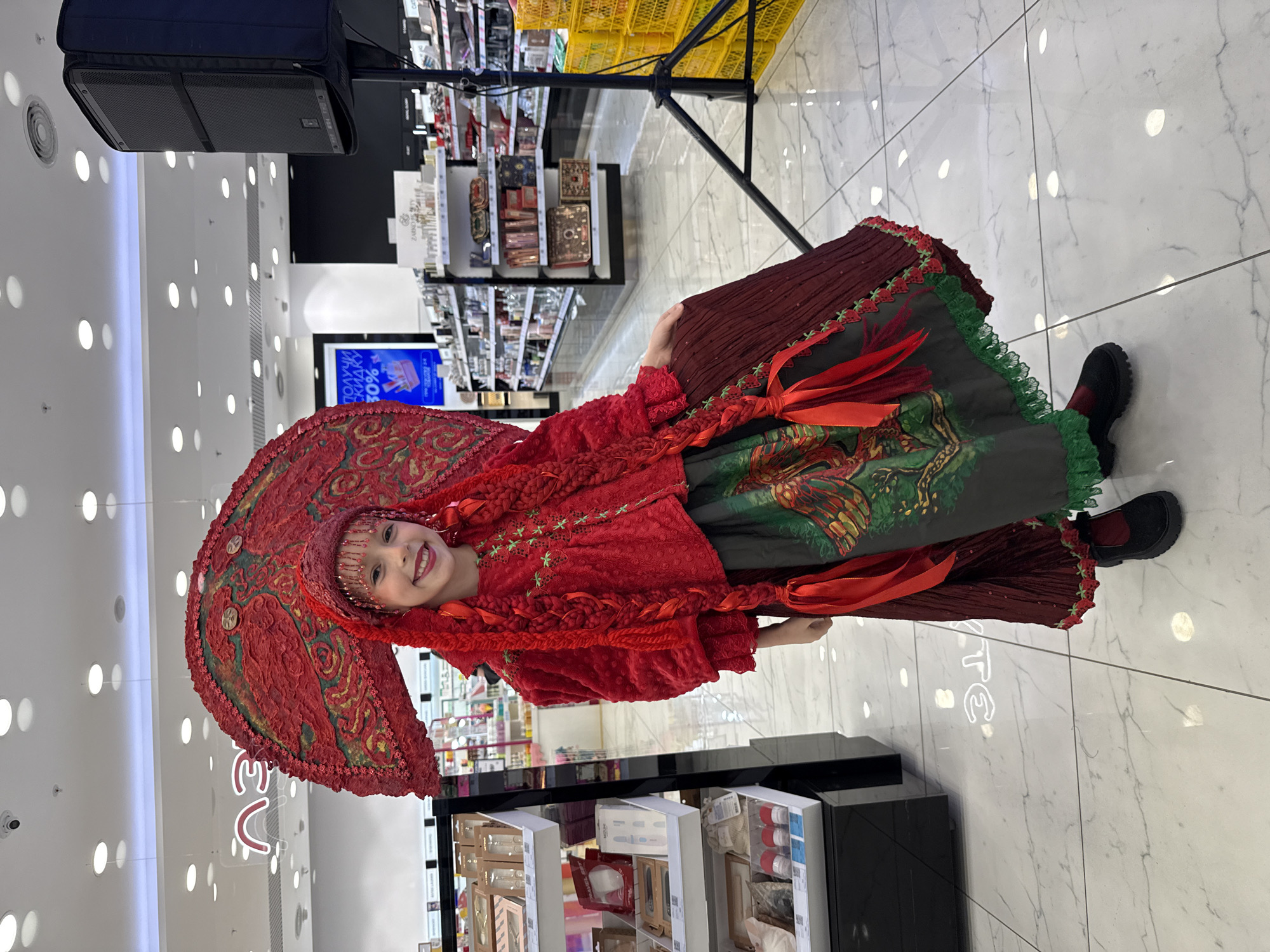 A young girl in traditional Russian attire smiles in what appears to be a department store or shopping mall. She is wearing a large, elaborate red headdress, a red top adorned with floral details, and a long skirt with green and red elements. Her braids are also decorated with red ribbons.
The background reveals shelves filled with various products, indicating a retail environment. The floor is light-colored with a marble-like pattern. Lighting fixtures are visible on the ceiling. There is also a display unit with makeup samples visible in the lower part of the image.
The setting suggests the image was taken in an urban commercial area, possibly Vladivostok, Russia.
