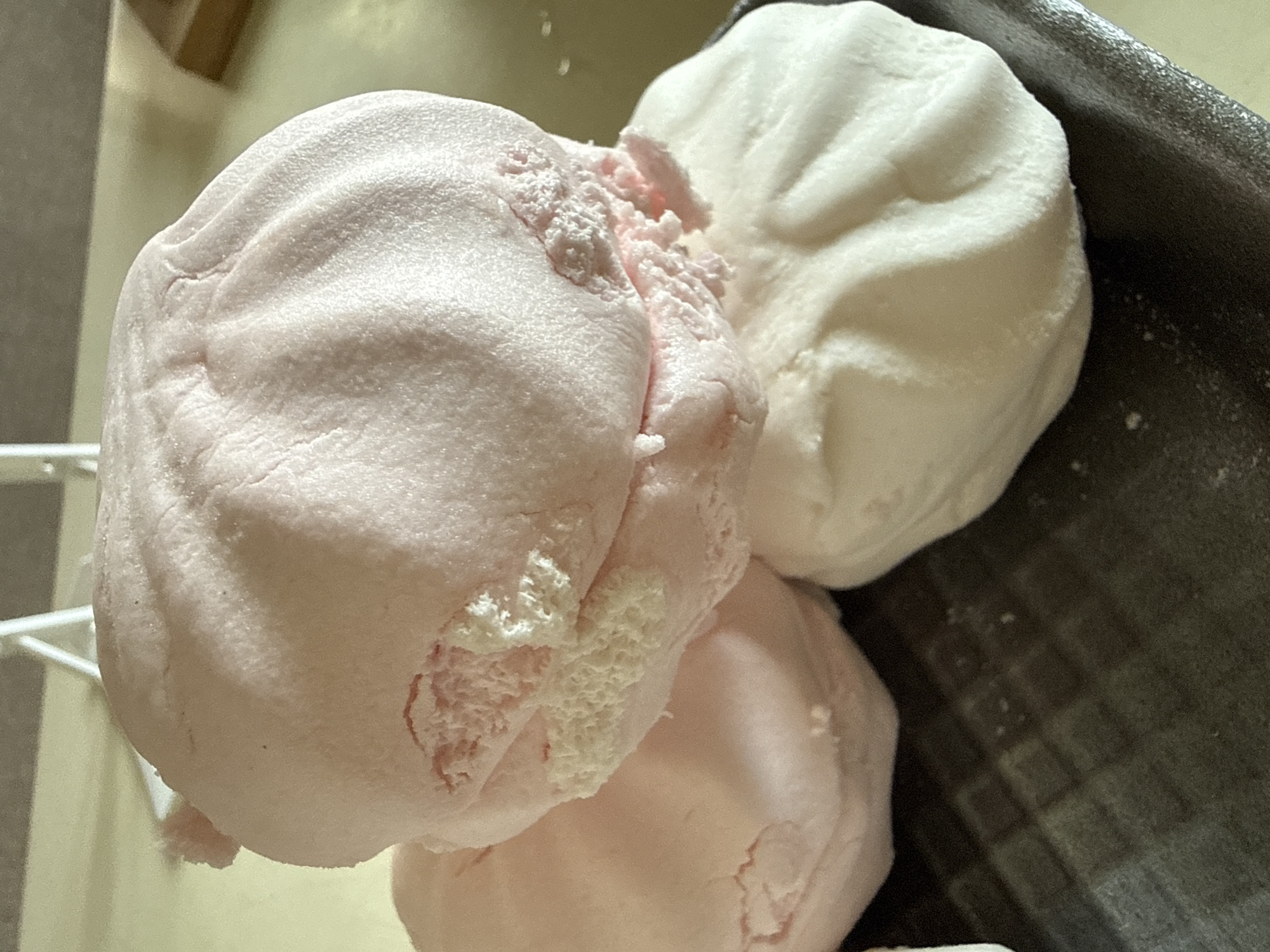 The image shows a close-up of three stacked, pastel-colored meringues. The largest meringue, on top, is pink and appears somewhat crushed, revealing a fluffy white interior. It has a ridged texture. Beneath it are two other meringues: one to the left, also pink and partially visible, and one to the right, which is white and has a smooth, slightly rippled surface. The meringues are resting on a dark, textured surface, possibly a styrofoam tray. The background is softly blurred, with a light-colored surface and hints of furniture or structures. No specific location in Okhtyrka, Ukraine, can be identified from this image.