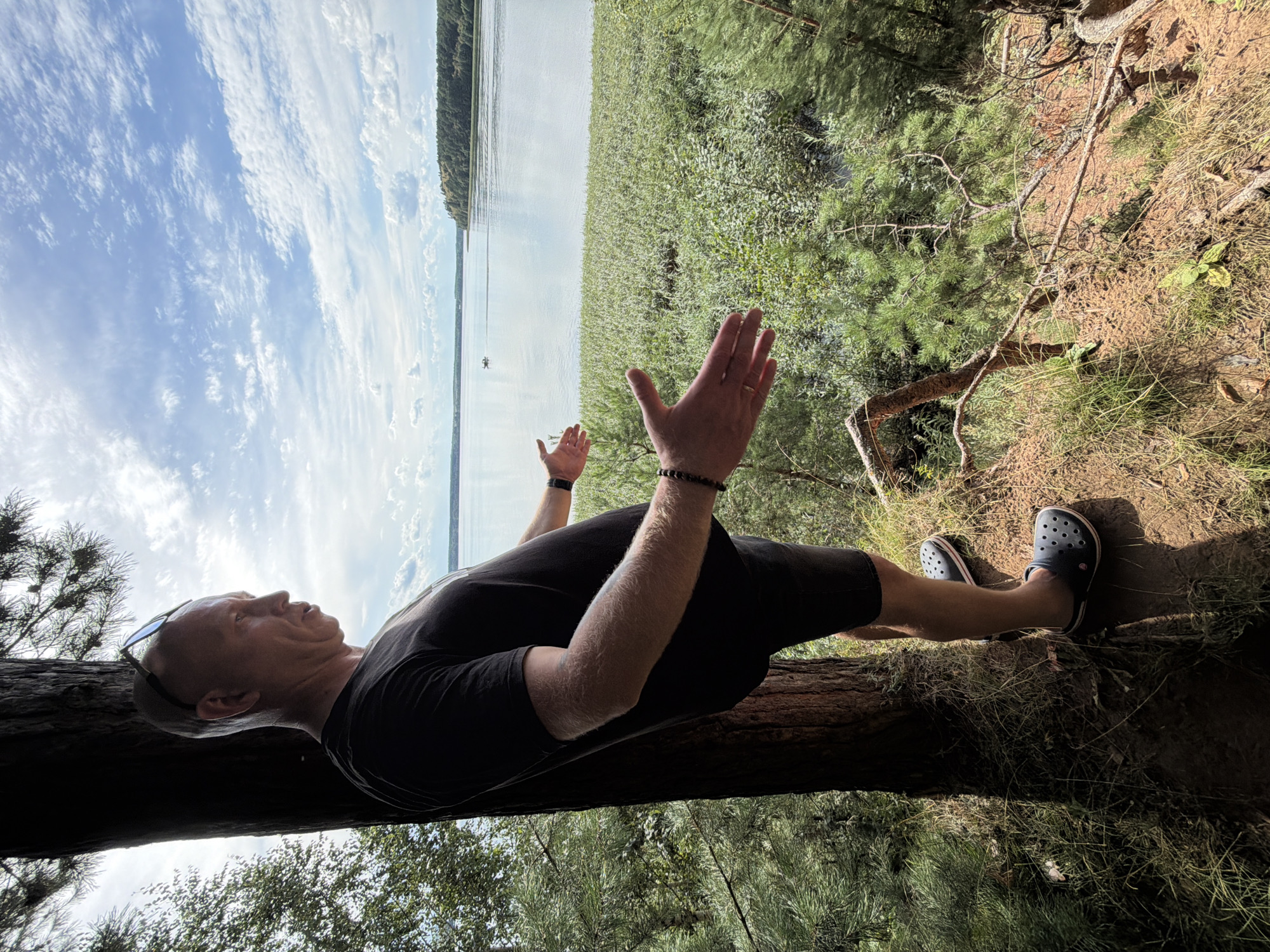 A bald, middle-aged man wearing a black t-shirt and black shorts, and dark clogs, stands near a tree on a grassy, wooded incline overlooking a large body of water. The man is facing left and has his arms outstretched, palms open. He is wearing sunglasses on his head and a bracelet on his left wrist. The water in front of him is calm and reflects the blue sky and white clouds. There is a small boat visible on the water in the distance, and a tree line on the far shore. The scene appears to be a natural landscape, likely a park or recreational area. The location is Votkinsk, Russia.