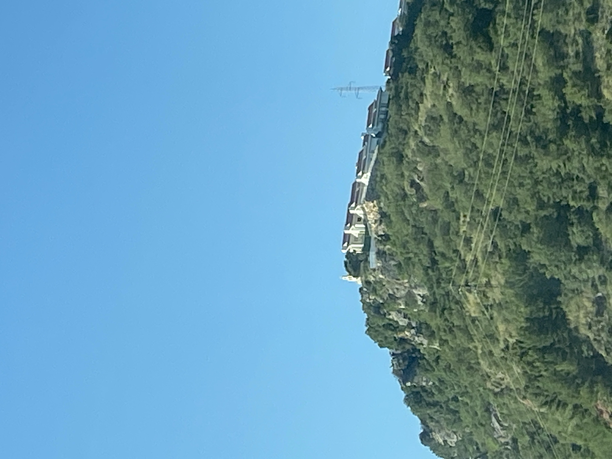 The image shows a hillside in Blizikuće, Montenegro, on a clear day. The hillside is covered in green vegetation and has buildings constructed along its slope. Power lines run along the side of the hill, and an electrical tower is visible in the distance. The sky is clear blue.