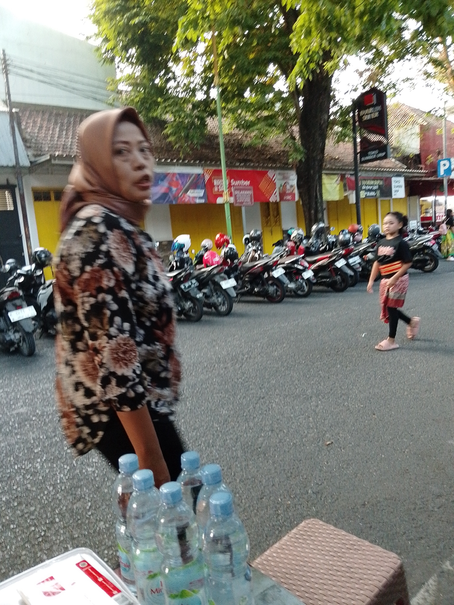 In Kecamatan Pati, Indonesia, a woman wearing a brown hijab and a patterned shirt stands in the foreground, looking to her left. Her shirt has a floral pattern with brown, white, and black colors. To her right, a young girl in a black shirt and a red and white patterned sarong walks on the asphalt road, looking forward. In the background, several motorcycles are parked in a row. The environment appears to be a street scene with buildings and trees visible. Several plastic bottles are arranged on a table in the lower foreground.