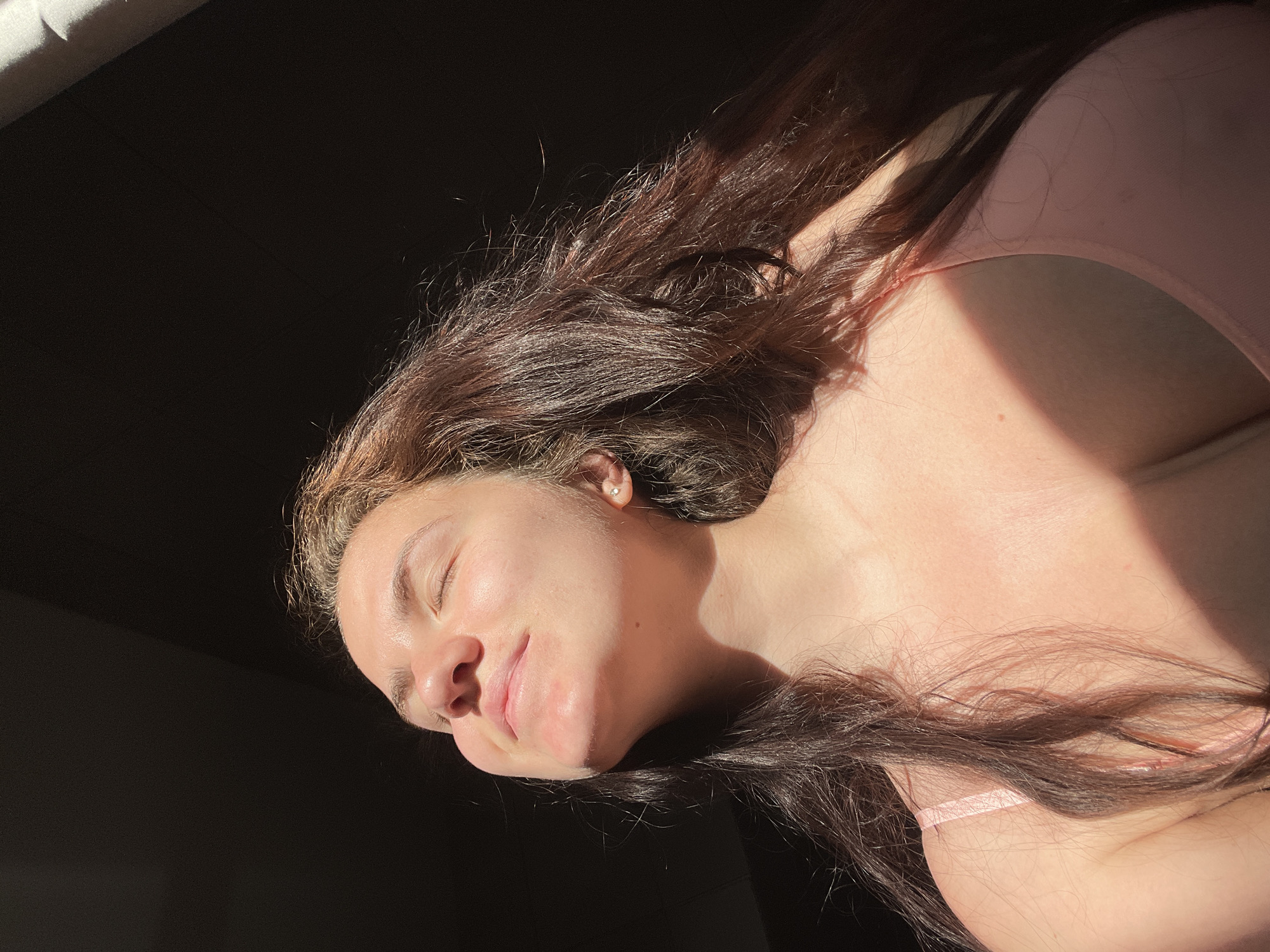 A woman is lying down. She has brown hair and is wearing a pink top. She appears to be resting with her eyes closed. The setting is unknown.