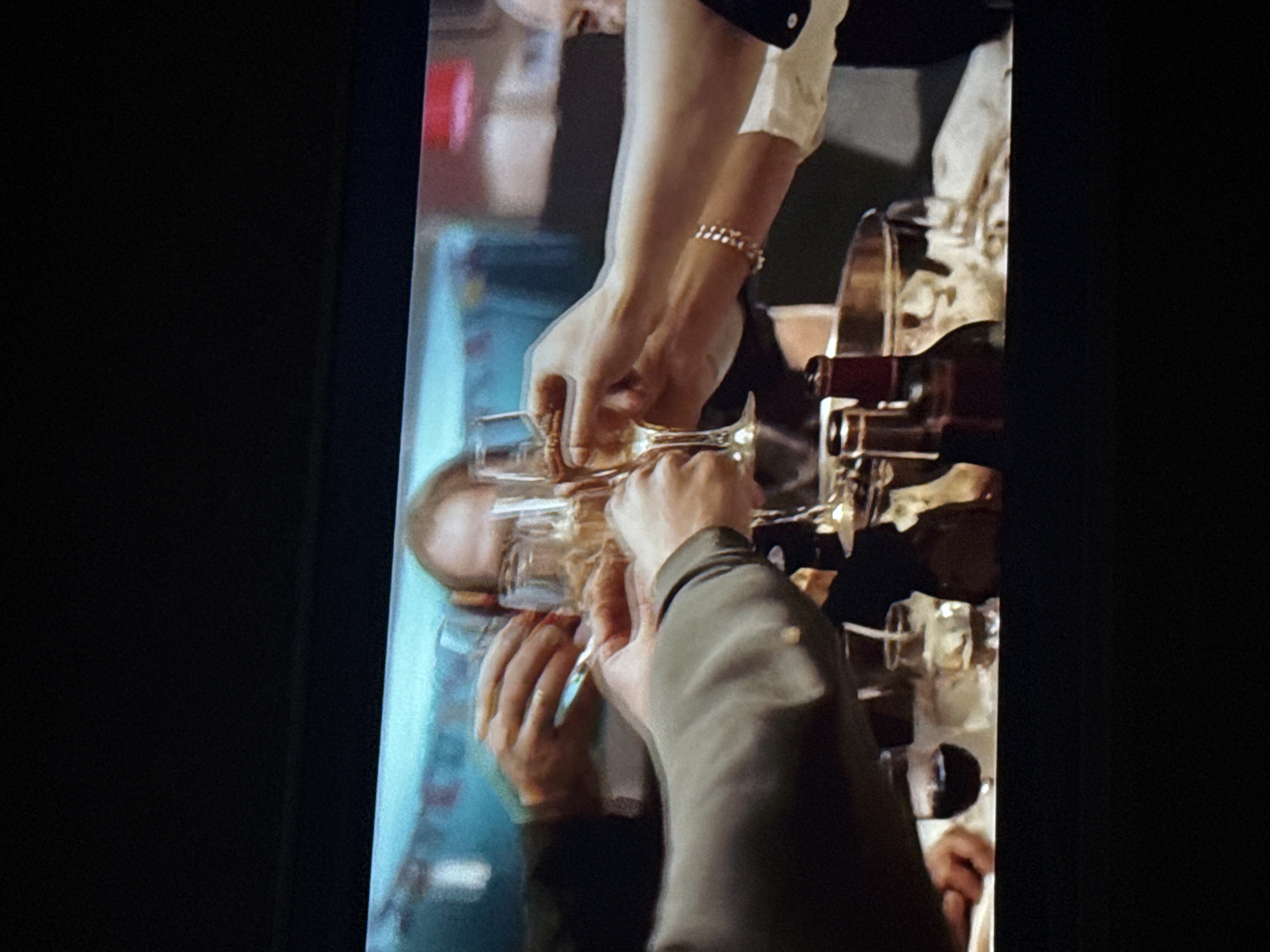 The image shows two people clinking champagne glasses. One person is wearing a white shirt with black trim and a bracelet. The other is wearing a dark gray jacket. A bottle of champagne is being poured into the glasses. The background is blurred, but appears to be a restaurant or bar. The location in the city of Mytishchi, Russia cannot be determined from the image.
