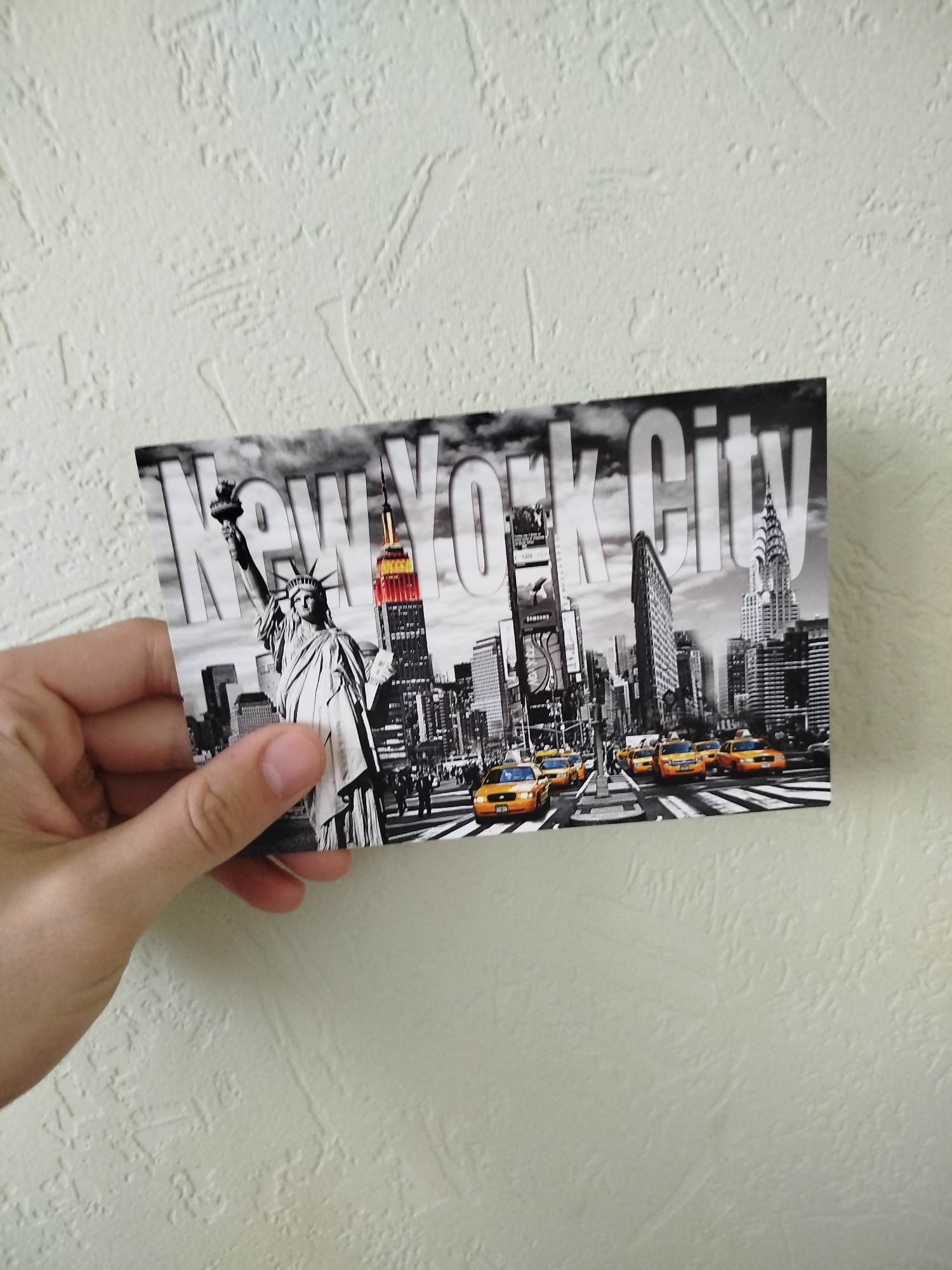 A postcard depicting New York City is held in a person's hand against a white, textured wall. The postcard features the Statue of Liberty, the Empire State Building, Times Square, and yellow taxis. The words "New York City" are printed across the top of the image. There is no indication of Varna, Bulgaria in the picture.