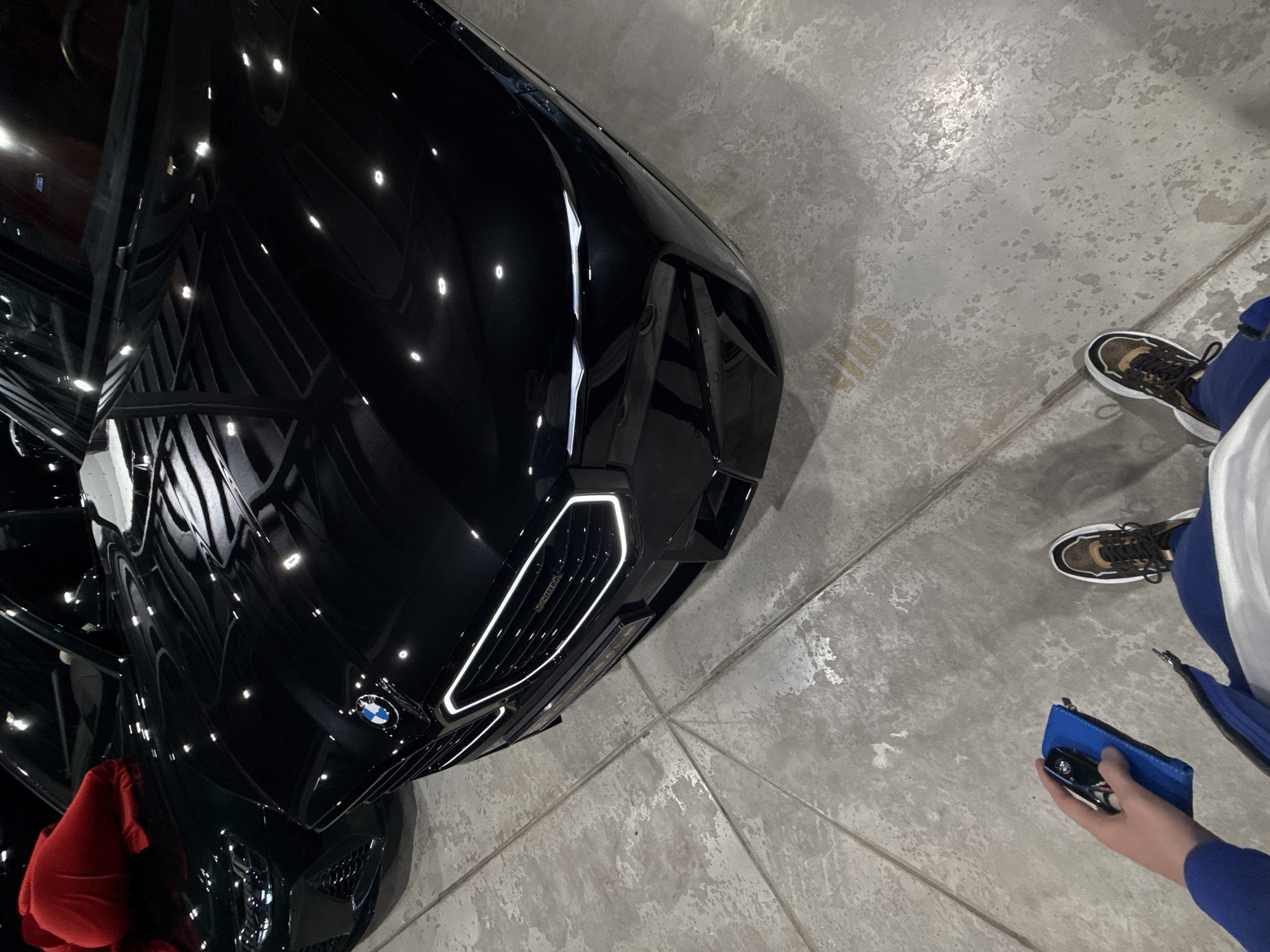 The image shows the front end of a black BMW car, likely parked on a gray concrete floor. The car is angled diagonally, with the front grille and BMW logo visible. Next to the car, to the right, are a pair of feet wearing sneakers with brown and black detailing. A hand is holding a small blue case that contains a toy car. The setting appears to be indoors, possibly a showroom or garage. The scene does not provide any information to suggest this is located in the city of Plovdiv, Bulgaria.
