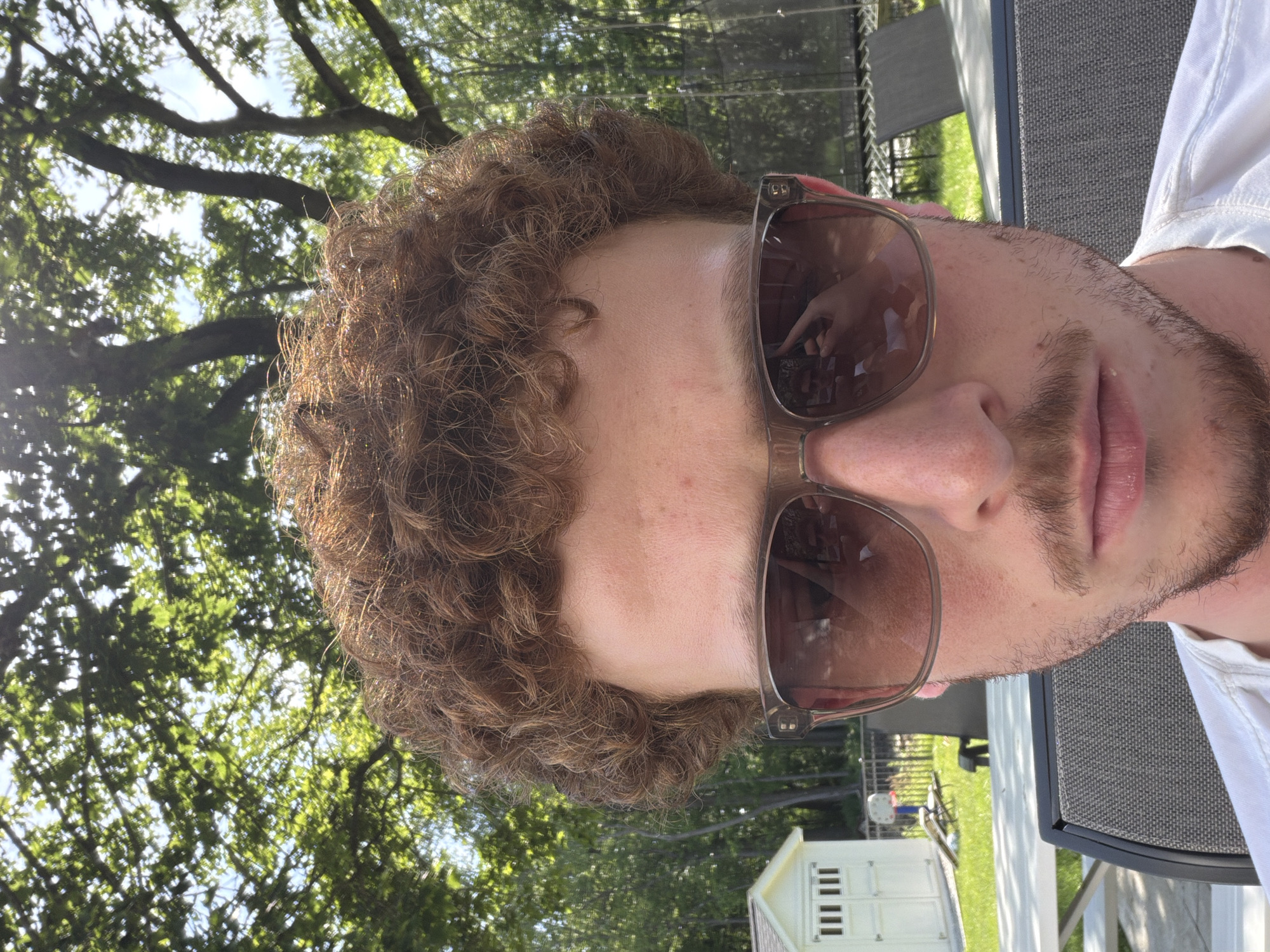 This rotated close-up portrait features a young man outdoors in what appears to be a residential backyard in Pepper Pike, United States of America. The subject, with curly brown hair and light stubble, is seen from the chest up, wearing brown-tinted sunglasses with clear frames and a white shirt. He looks towards the viewer with a neutral expression.
The background is dominated by lush green trees and foliage, indicating a bright, sunny daytime environment. Reflections in the sunglasses show a hand, possibly the subject's, holding a device, suggesting a selfie or being photographed. To the left side of the frame, a small white shed with multiple small rectangular windows is visible in the mid-ground. To the right, outdoor furniture, including a grey mesh chair and a table, and a dark fence are present. The strong sunlight casts highlights on his hair and illuminates the scene, consistent with a clear, pleasant day. No visible text is present in the image.