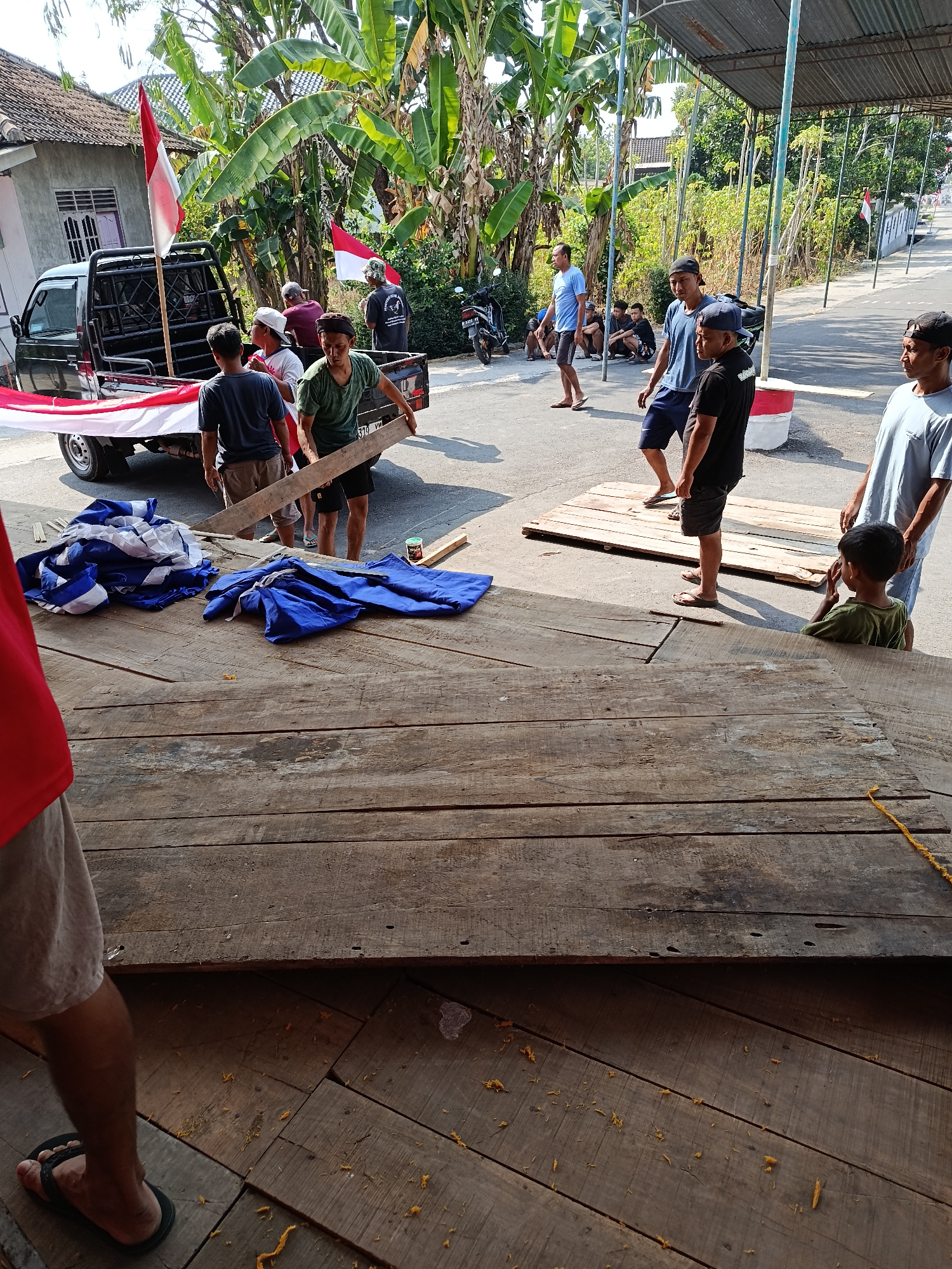 The image depicts an outdoor scene in Jurug, Indonesia, likely during daylight. In the foreground, a large wooden platform is visible, with several planks showing signs of wear and tear. To the left, the lower body of a person wearing a red shirt and light-colored shorts is present, standing on wooden planks.
In the middle ground, a black pickup truck is parked, adorned with a red and white Indonesian flag. Several men are engaged in an activity around the truck. One man in a green shirt and black shorts is carrying a wooden plank, assisted by another man. Other individuals are standing or sitting nearby, some observing the activity. Folded blue and white fabric, possibly banners or flags, are placed on the ground next to the truck.
Further in the background, a street is visible with a few motorcycles and more people. Banana trees and other lush vegetation surround the area. The overall environment suggests a rural or semi-urban setting, possibly preparing for an event or celebration given the presence of the Indonesian flag. The scene appears to be bustling with activity as the individuals work together.