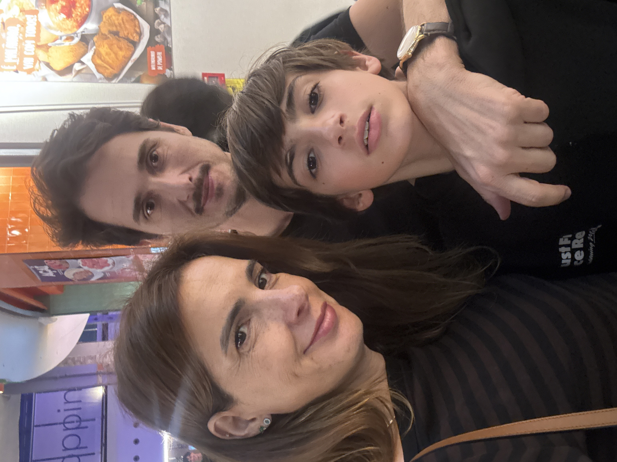 The image shows a selfie taken indoors of a family of three. The mother is in the foreground, smiling at the camera. Behind her is the father, also looking at the camera, slightly to the left of the frame. Between them is a child with short, dark hair and a hand with a watch resting near their neck.
The background suggests they are in a public space, possibly a restaurant or shopping area, in Vila Nova de Gaia, Portugal. There's a menu poster visible above the father's head with the text "É MALANDAO, VAI COM TODOS!" and an advertisement for "DE POMBAL". A sign with the partial word "ppin" is visible above the mother. The woman is wearing a striped dark top and a strap of a purse is also visible. The child and the father are wearing black shirts. The T-shirt has some words that partially reads "ust Fi ice Re".