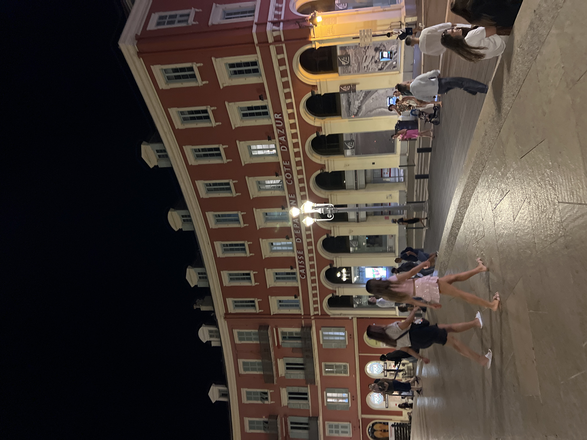 The image captures a night scene in Place Masséna, Nice, France. A grand, salmon-pink building with white trim and numerous windows lines the background. Above the arched ground floor, white lettering spells out "CAISSE D'EPARGNE COTE D'AZUR." Several streetlights illuminate the paved area where people are walking and standing. In the foreground, two women are walking from left to right. The woman on the left wears a dark skirt and a white top, while the woman on the right is in a pink sequined skirt and a light-colored top. Other individuals are scattered throughout the scene, including a group sitting on a lower level to the right, and others standing or walking near the building. The overall atmosphere is lively and urban, with the warm glow of the lights reflecting on the wet pavement.