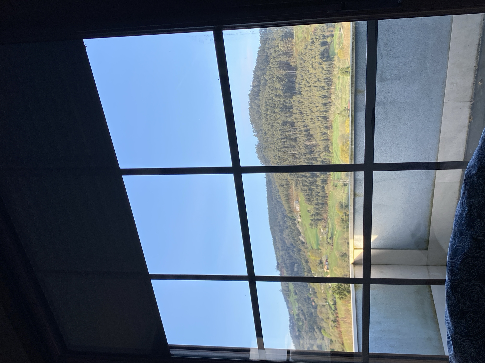 The image shows a view through a grid-patterned window, likely a skylight, looking out onto a landscape. The landscape includes a densely forested hillside with patches of green fields and some buildings in the distance. Part of the view also includes an adjacent gray concrete balcony or wall. A dark-colored shade or panel is partially obscuring the upper left portion of the window. A patterned curtain partially covers the right side of the window. The sky is blue and visible through some of the window panes. The location is identified as Villaviciosa, Spain.