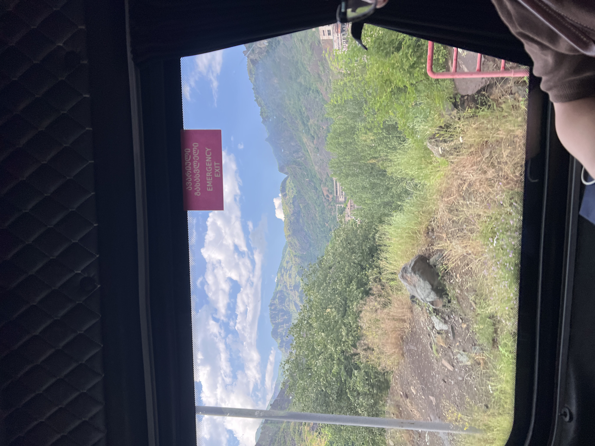 The image, captured from the interior of a vehicle, looks out through a window at a mountainous landscape. A pink sign with Georgian script and the words "EMERGENCY EXIT" is affixed to the upper part of the window. Below the sign, the view reveals lush green trees and dry grass covering the slopes of rugged mountains under a sky dotted with white clouds. In the distance, structures suggestive of buildings and a castle or fortification are visible on the mountain peaks. The foreground of the scene includes dry vegetation, rocks, and a portion of a metal railing. The lower edge of the window frame and a person's arm in a brown shirt are also visible. This scenery is consistent with the natural environment surrounding Alaverdi, Armenia, a city known for its mountainous terrain and historic monasteries.