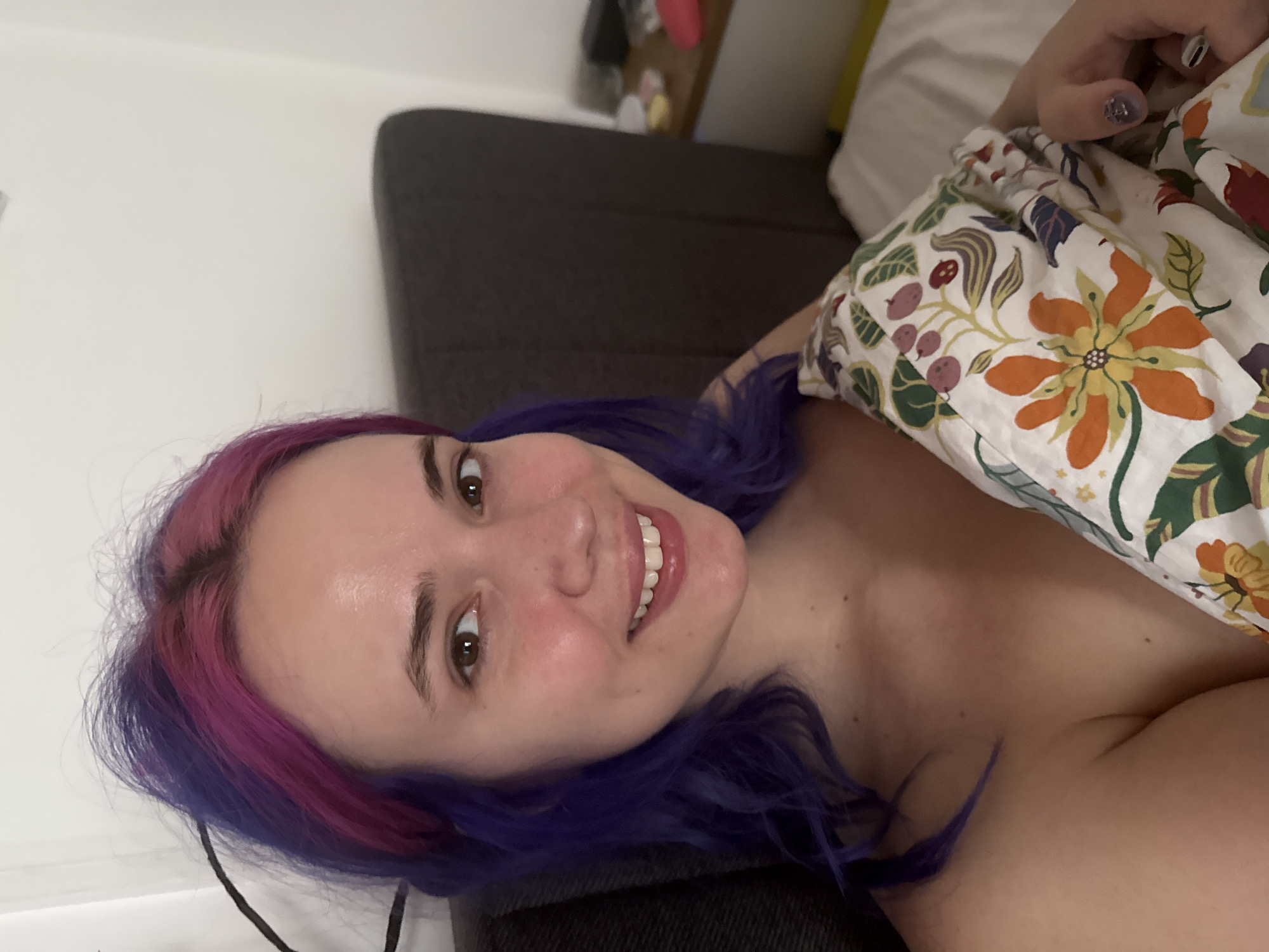 A smiling young woman with distinctive two-toned hair, featuring bright pink roots transitioning to vibrant purple, is captured in a close-up selfie. Her brown eyes look directly at the camera, and her face is bright with a wide, natural smile, revealing white teeth. Her cheeks show a slight natural flush.
She is depicted indoors, likely reclining in bed or on a sofa, with her upper body partially visible. A colorful blanket or duvet with a prominent floral and botanical pattern covers part of her torso. The pattern features large orange flowers, green leaves, and reddish-purple berries against a white background. Her hand, with fingernails painted in a light purple glittery polish, rests on the patterned fabric.
Behind her, a dark gray or black headboard or backrest of furniture is visible, juxtaposed against a plain white wall. The overall impression is one of relaxation and happiness. The image appears to be taken in good indoor lighting, suggesting either daytime or a well-lit interior. There is no visible text in the image. The provided context indicates the location is the City of Zagreb, Croatia.