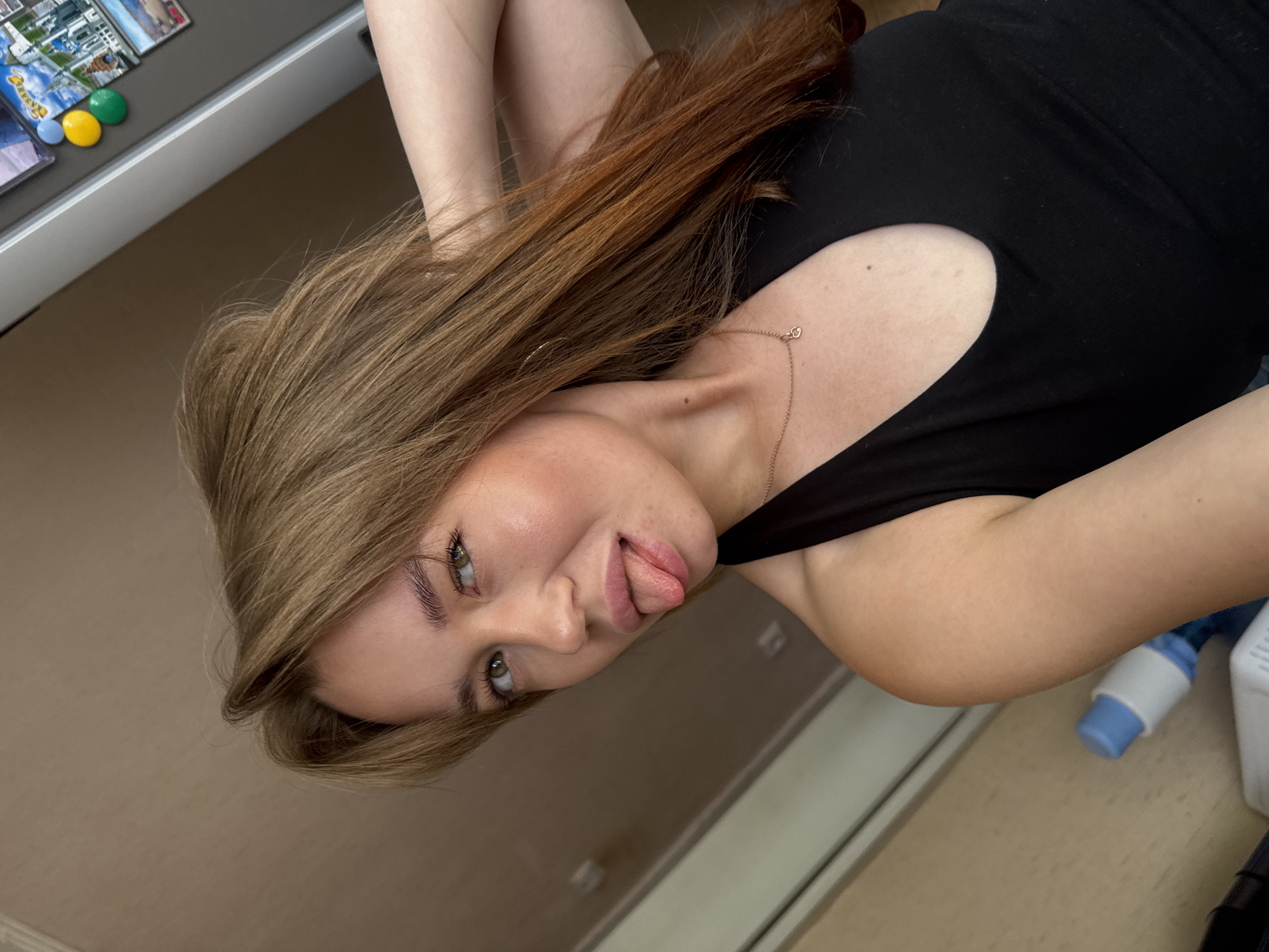 In a selfie taken indoors, a young woman with light brown hair, styled with bangs, is shown from the chest up. She has light green eyes and is sticking her tongue out, with her left hand behind her head. She is wearing a black tank top with a V-neck and a delicate rose gold necklace with a heart pendant. A mole is visible on her neck. The background appears to be a domestic setting, with a wall and a refrigerator adorned with magnets. The scene is set in Chaikovsky, Russia, a city known for its rich cultural heritage and natural beauty, nestled along the Kama River.
