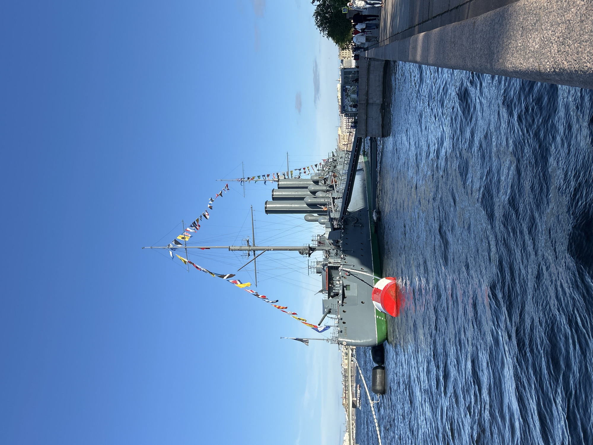 This image captures a bright, clear day with a deep blue sky over a body of water, likely a river or canal. A large, grey warship, adorned with a string of colorful nautical flags, is moored alongside a stone embankment. The ship has multiple smokestacks and a prominent gun turret. Mooring buoys, one red and white and others black, are visible in the water near the ship. On the embankment, a paved walkway runs parallel to the water, with several people walking along it. In the background, buildings and a bridge are visible, suggesting an urban setting. The overall scene is one of a historical naval vessel in a metropolitan harbor on a pleasant day.