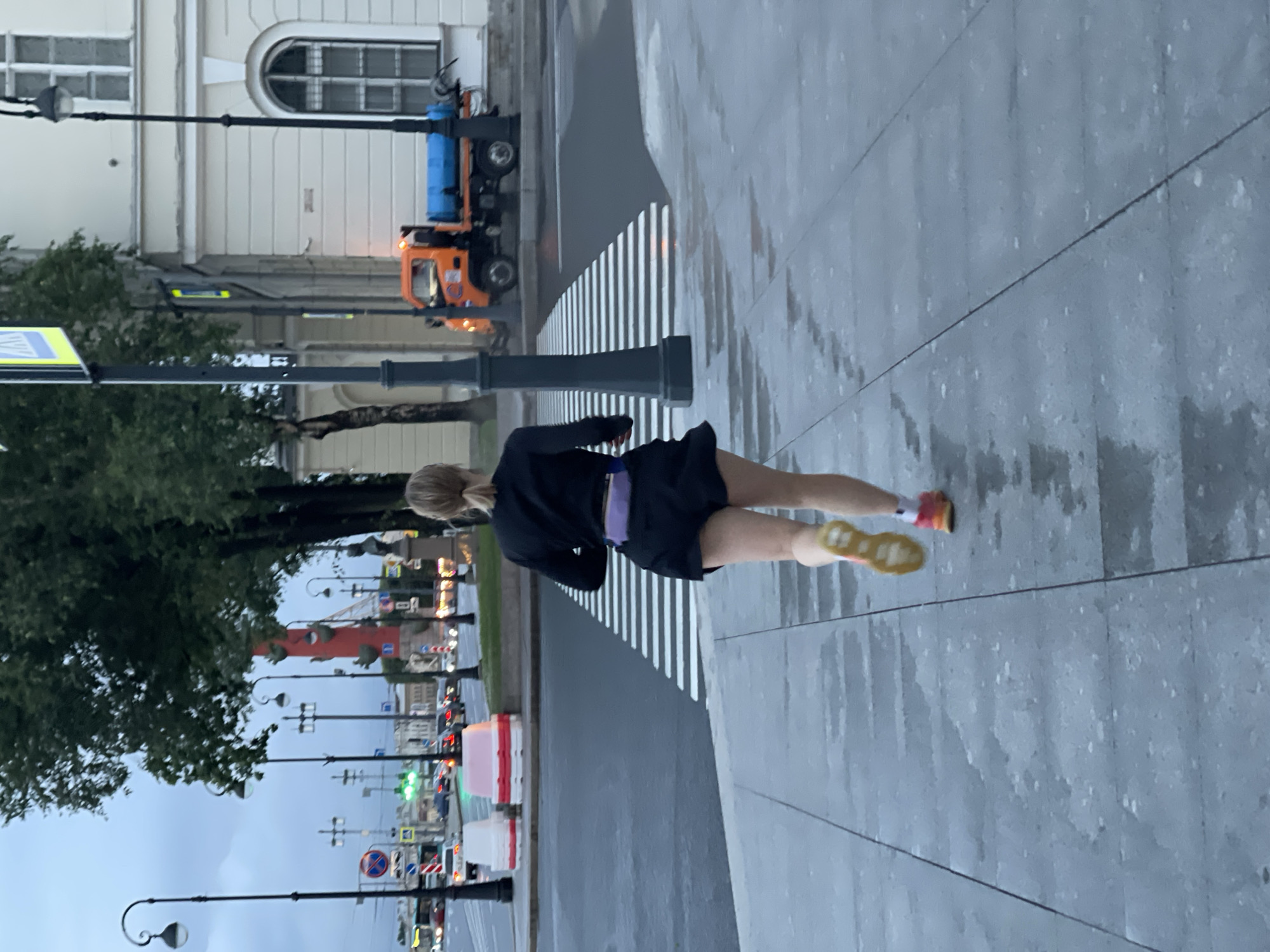 A lone female runner is captured from behind, mid-stride, on a wet sidewalk in Saint Petersburg, Russia. She is wearing a black t-shirt, black shorts, and bright athletic shoes. A purple fanny pack is visible around her waist. The runner is approaching a crosswalk with white stripes on a wet asphalt road.
In the background, a bright orange maintenance truck with a blue tank is parked. Several streetlights with decorative lamps are present. A tall, thin red structure, possibly a monument or a column, stands out against the overcast sky. The architecture of the buildings suggests a European city, consistent with Saint Petersburg. The overall atmosphere is somewhat muted due to the cloudy weather and the wet conditions, likely indicating recent rain. The image appears to have been taken during the day, but the lighting suggests it might be early morning or late afternoon.
