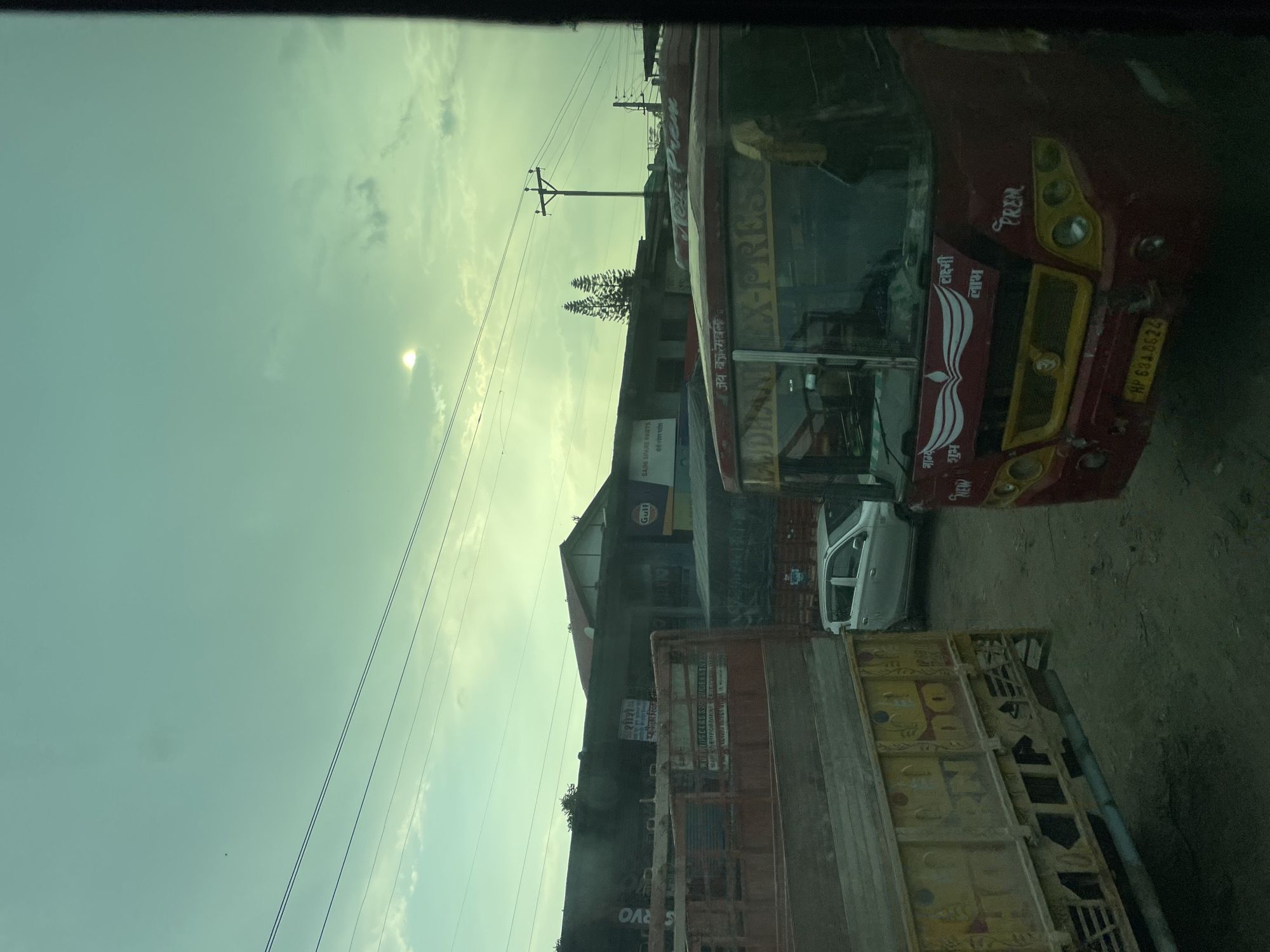 The scene depicts a street view in Gaggal, India, taken from inside a vehicle, possibly a bus or car. A red bus with yellow accents labeled "BAJDHANI EX-PRES" and "PREM" dominates the right side of the frame, partially obscured by the viewing angle. The bus has license plate "HP 634.8624". Adjacent to the bus is a white car. In front of that there is a truck with a yellow/brown cargo container. A Gulf gas station or similar structure is in the background, along with power lines. A building is labeled "SAINI SPARE PARTS". The sky is overcast, lending a slightly hazy quality to the scene.