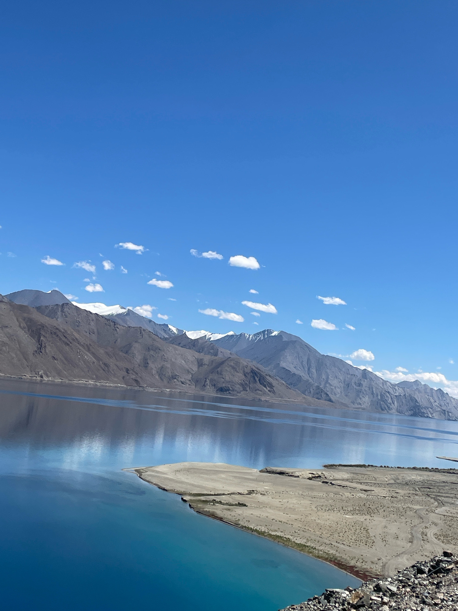 The image depicts a vast, tranquil lake with strikingly clear blue water reflecting the surrounding mountainous terrain and a bright blue sky dotted with scattered white clouds. The lake's edge in the foreground features a sandy shoreline with some rocky debris. In the background, a range of rugged, barren mountains stretches across the horizon, their peaks dusted with snow. The scene conveys a sense of serene, natural beauty in a remote, high-altitude environment, likely in Ladakh, India, given the characteristic landscape.